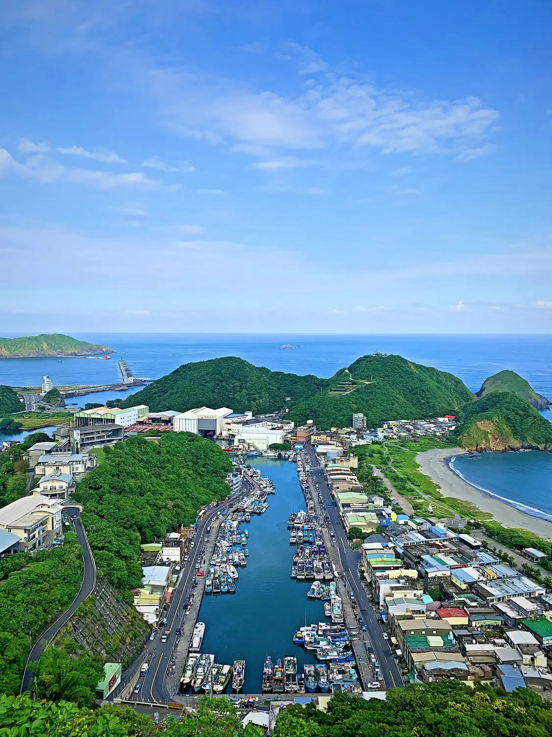 Aerial view of fishing harbor in Taiwan with rows of moored boats, coastal town, and green headlands meeting the blue ocean