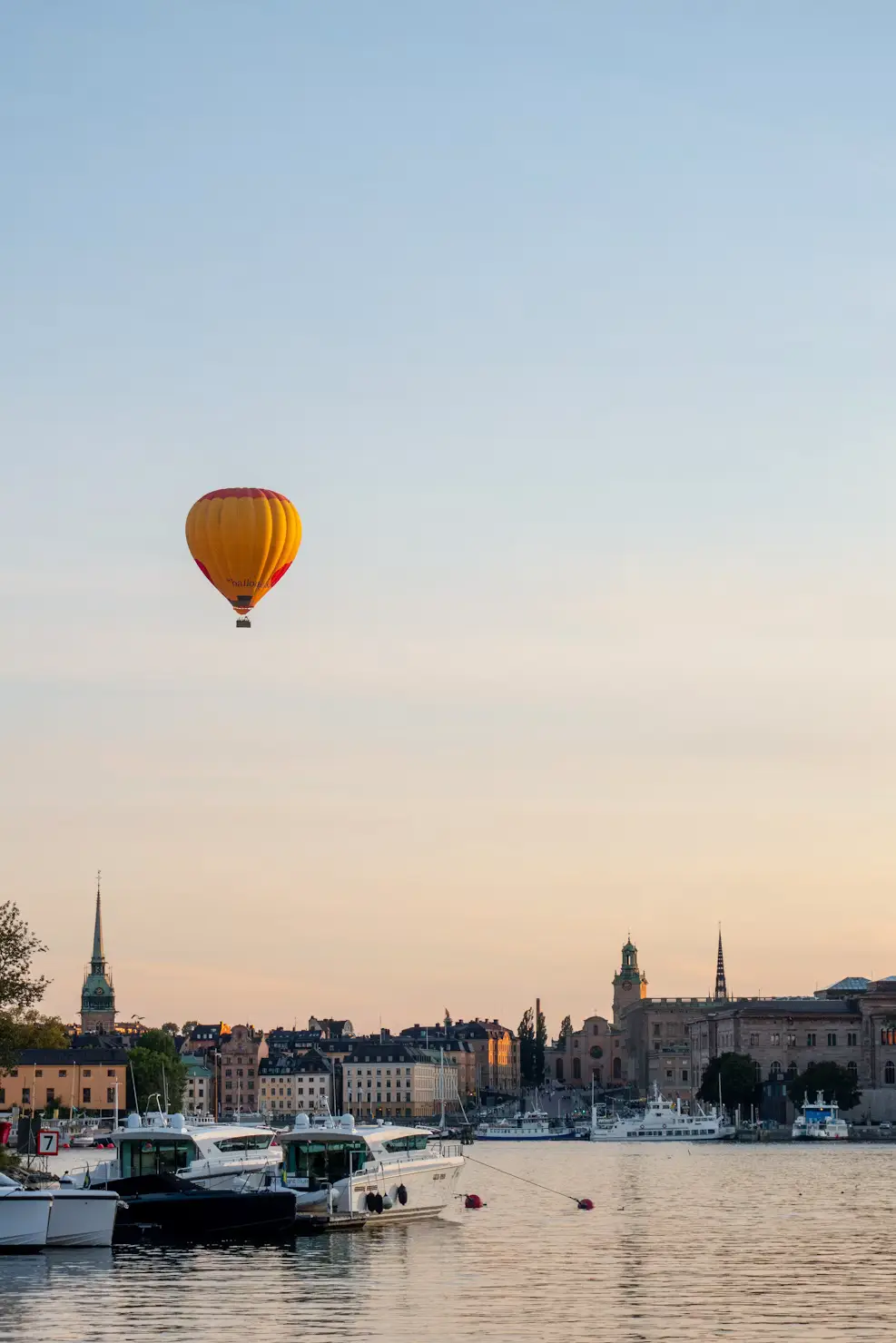 A hot air balloon floats over a waterfront city skyline at dusk, with church spires and boats visible along the harbor below.