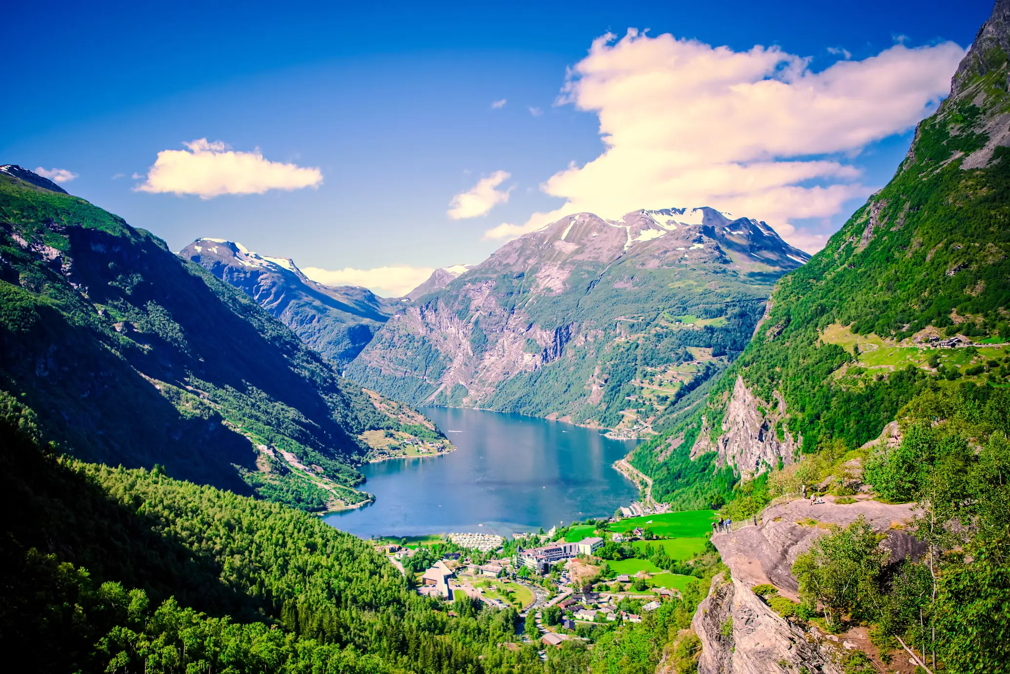 Sweeping view of Geirangerfjord from above, with green mountainsides plunging to the blue water and a village at the fjord's edge.