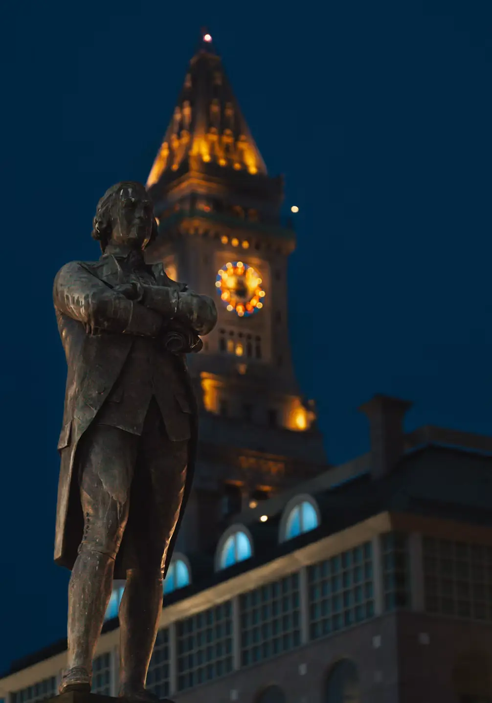 Bronze statue silhouetted at night against the glowing, illuminated clock tower of Boston's Custom House.