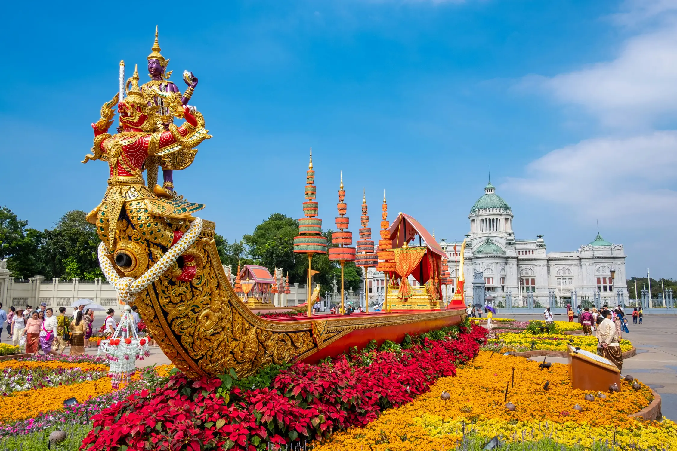 Elaborately decorated golden royal barge float with a mythical deity figurehead surrounded by red and yellow flower displays, with the Ananta Samakhom Throne Hall in the background, Bangkok, Thailand