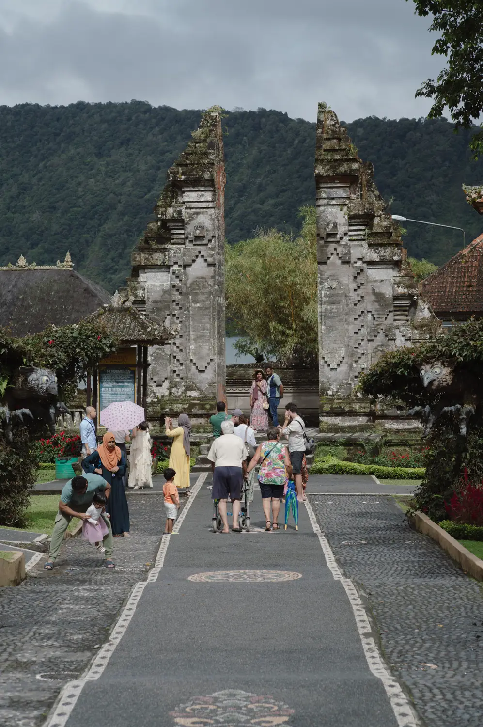 Visitors walking through a split gate (candi bentar) temple entrance along a stone pathway with mountains in the background in Bali, Indonesia.