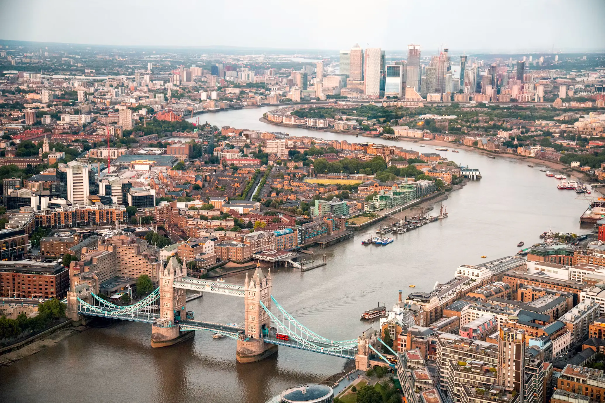 Aerial view of London showing Tower Bridge spanning the River Thames, with the city's dense rooftops stretching toward the Canary Wharf skyline in the distance.