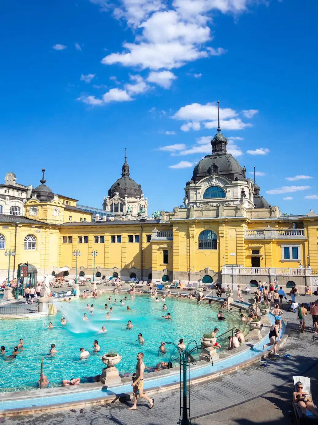 Large outdoor thermal pool crowded with bathers beside grand yellow historic buildings.