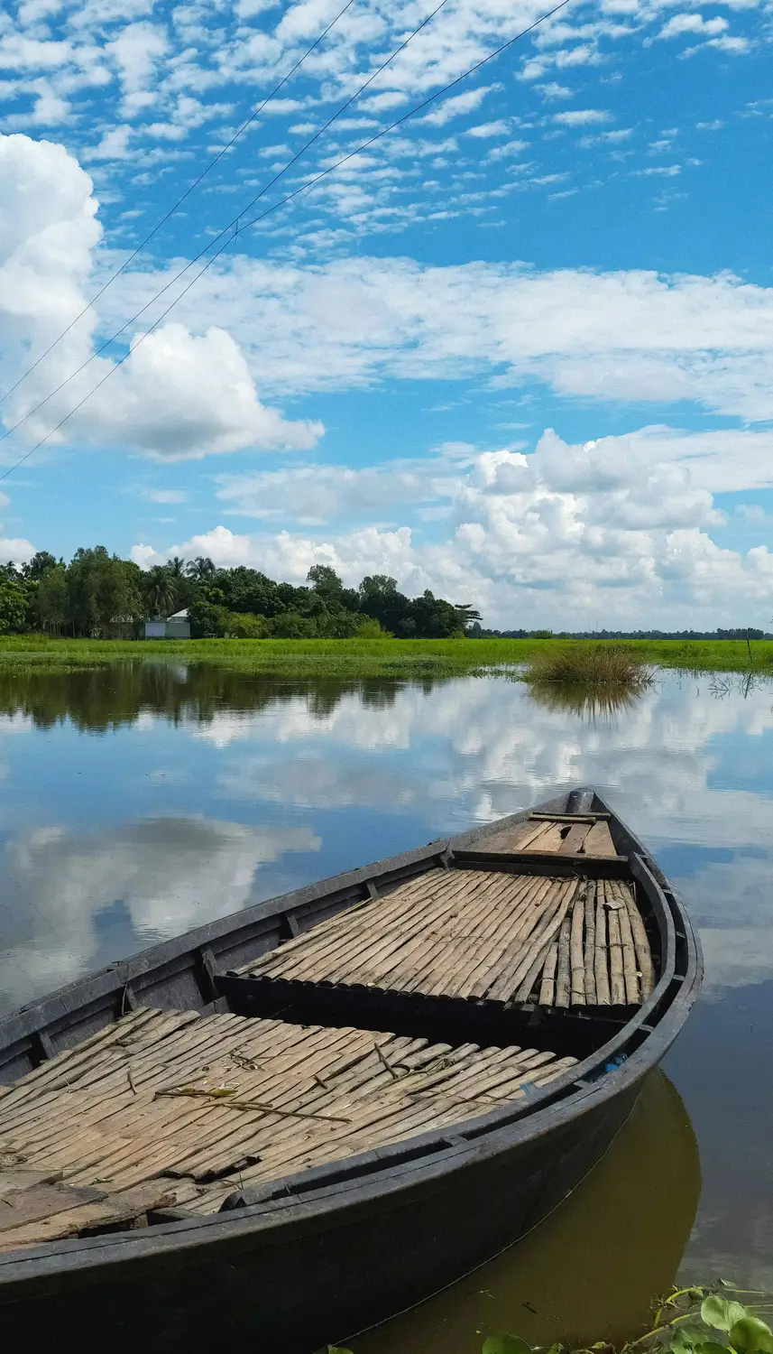 Traditional wooden boat moored on a calm flooded river with cloud reflections and green paddy fields, rural Bangladesh