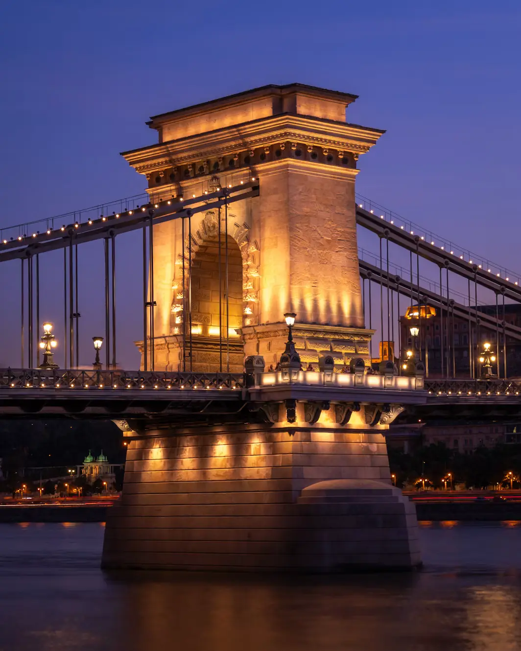 Illuminated suspension bridge tower at dusk, with warm lights reflecting on the river below.