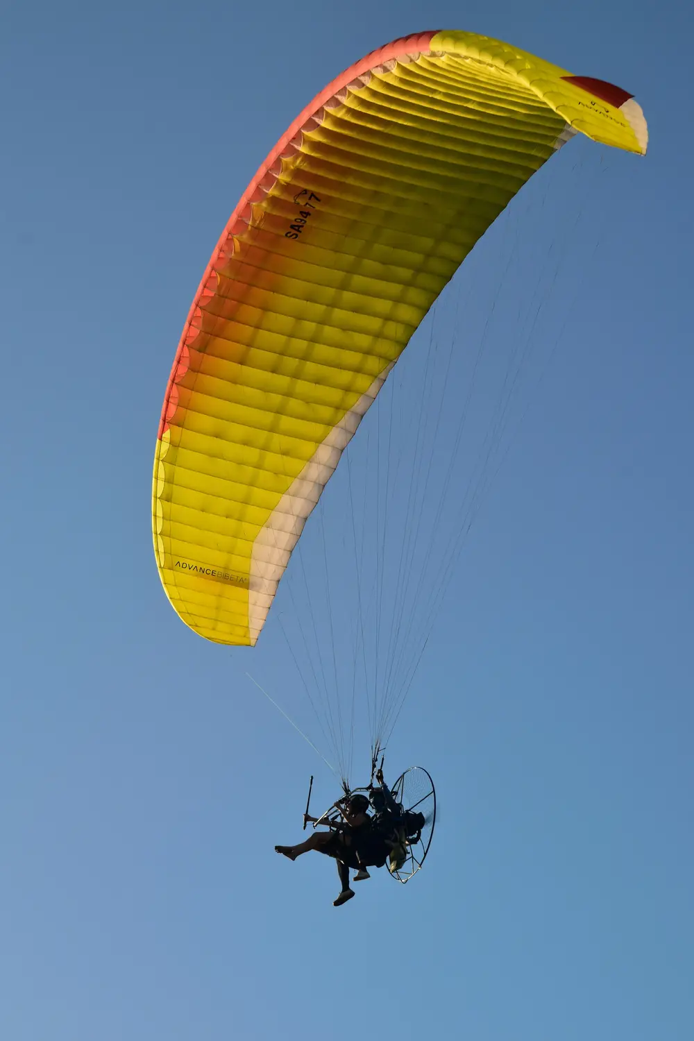 Tandem powered paraglider with a yellow and orange canopy flying against a clear blue sky in the Philippines.