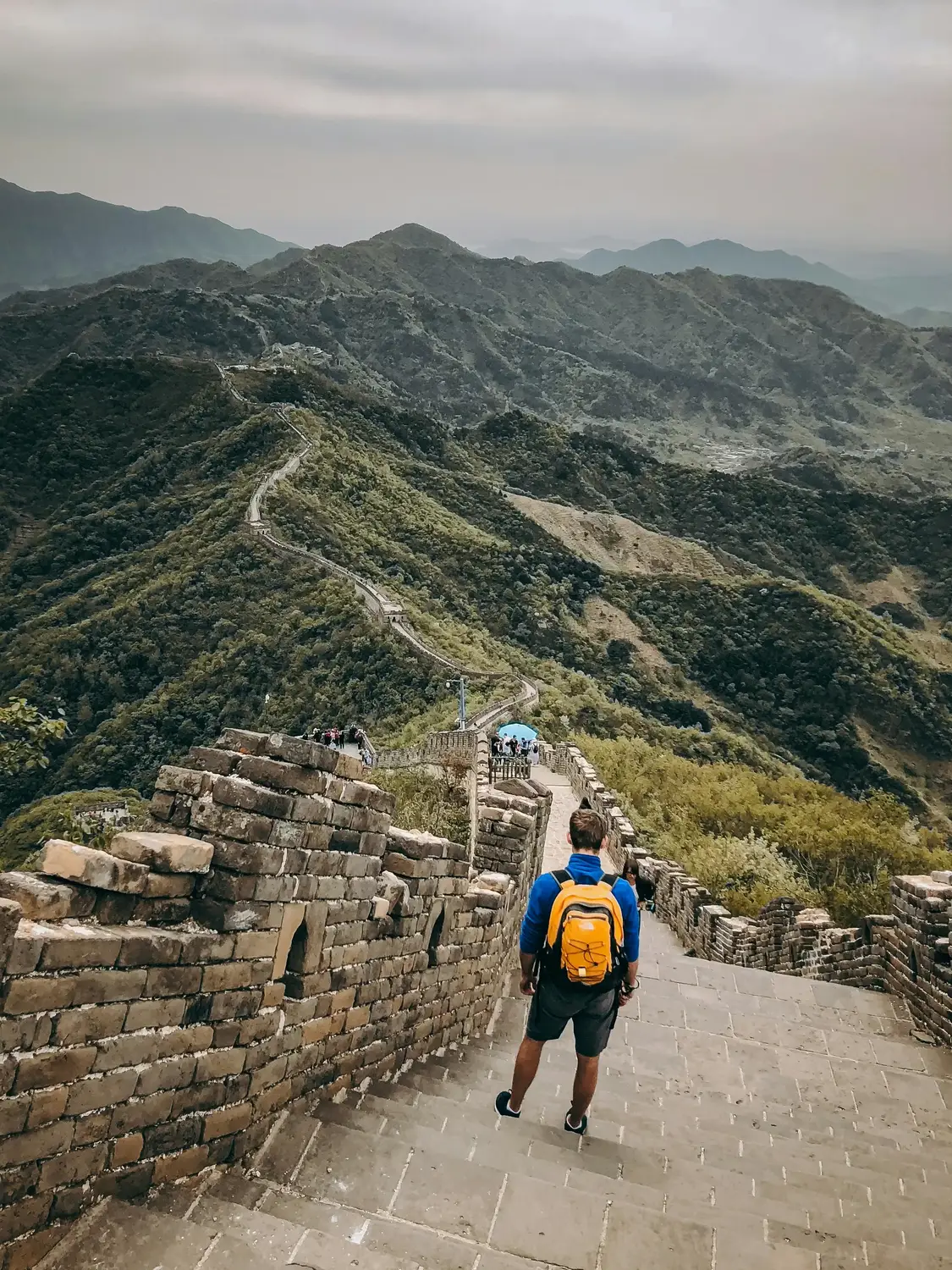 Hiker with yellow backpack walking down Great Wall of China steps with green mountain valley panorama