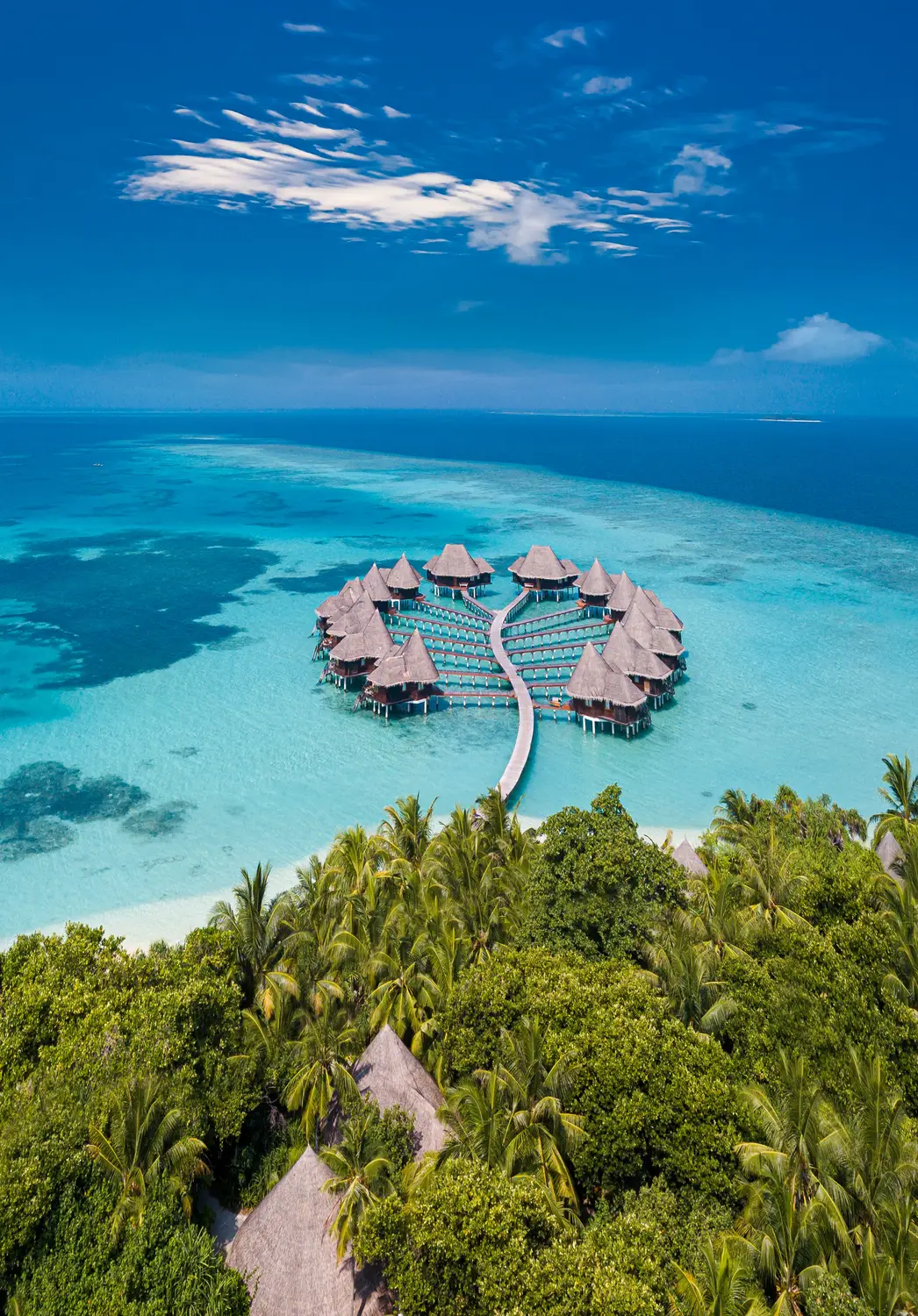 Aerial view of circular cluster of overwater bungalows connected by wooden jetty surrounded by turquoise lagoon and tropical palm trees in Maldives