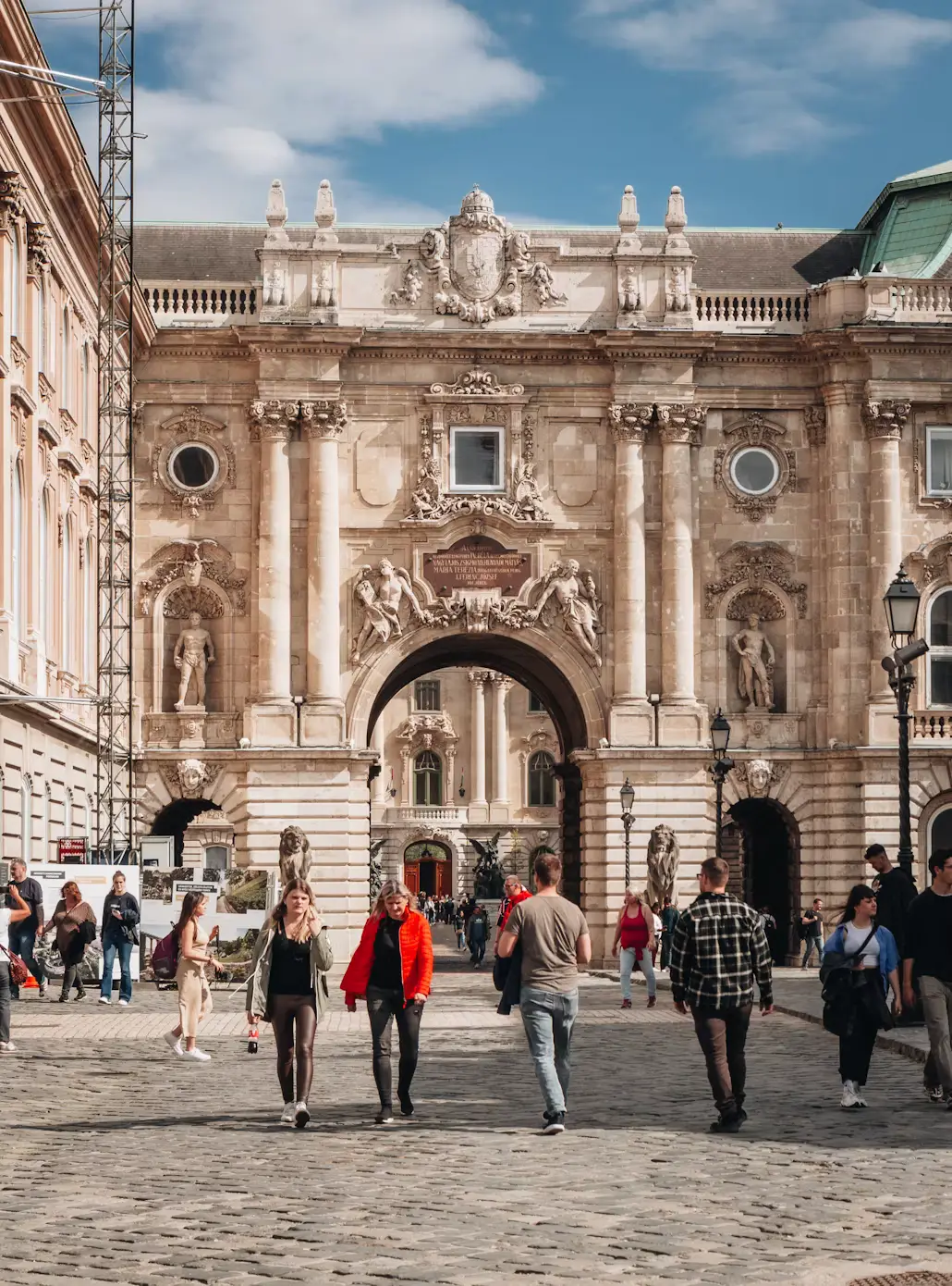 Ornate historic palace gate with statues and columns, with pedestrians walking through a cobblestone square.