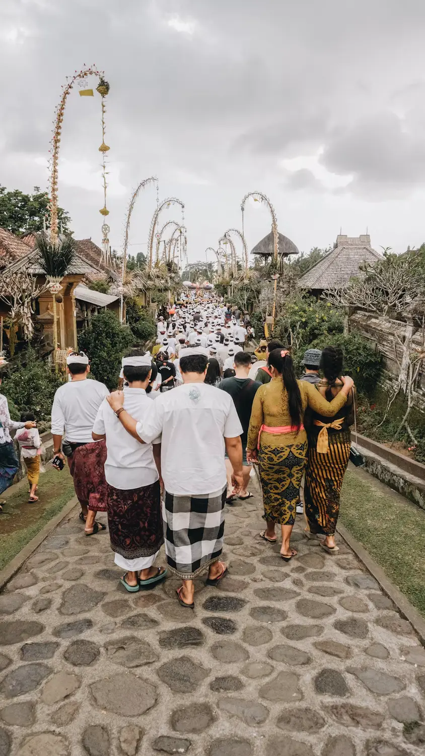 Balinese people in traditional attire walking in a temple procession along a stone path with penjor poles in Bali, Indonesia.