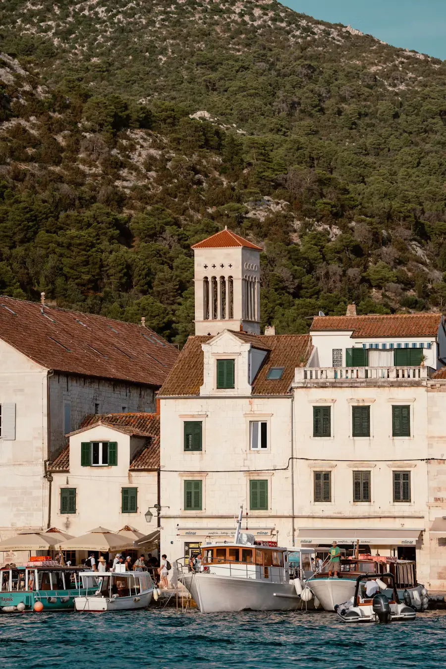 Waterfront buildings with a bell tower along a harbor, boats docked below and a green hillside rising behind, Croatia.