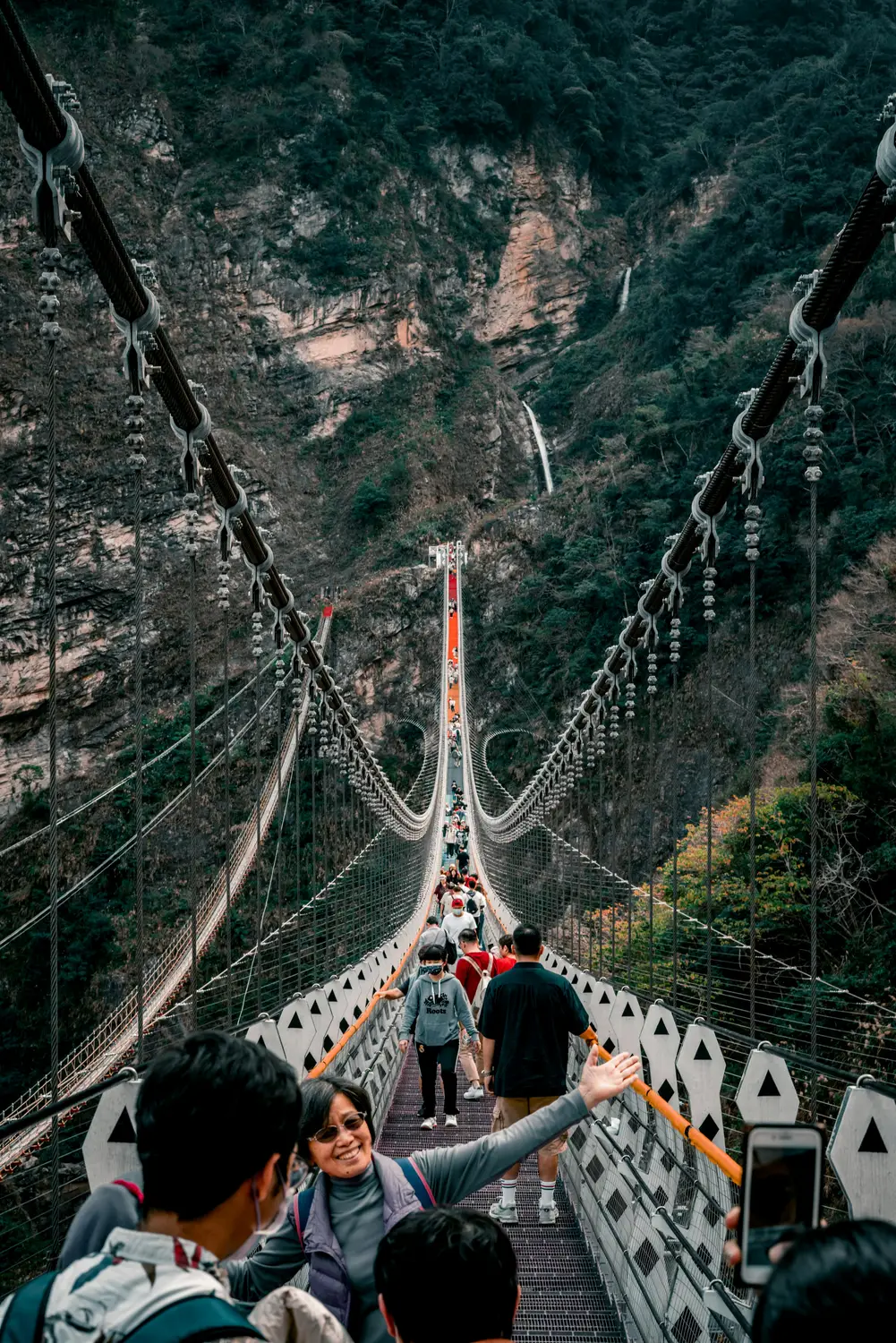 Visitors crossing a long suspension footbridge over a forested mountain gorge with a waterfall in Taiwan
