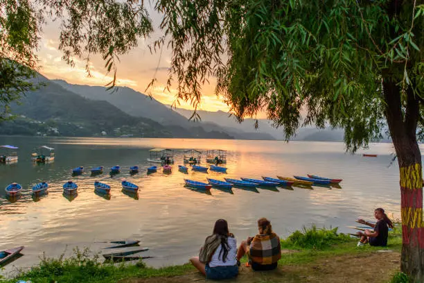 People relaxing by a lakeside at sunset with colorful boats and mountains, Phewa Lake, Pokhara, Nepal