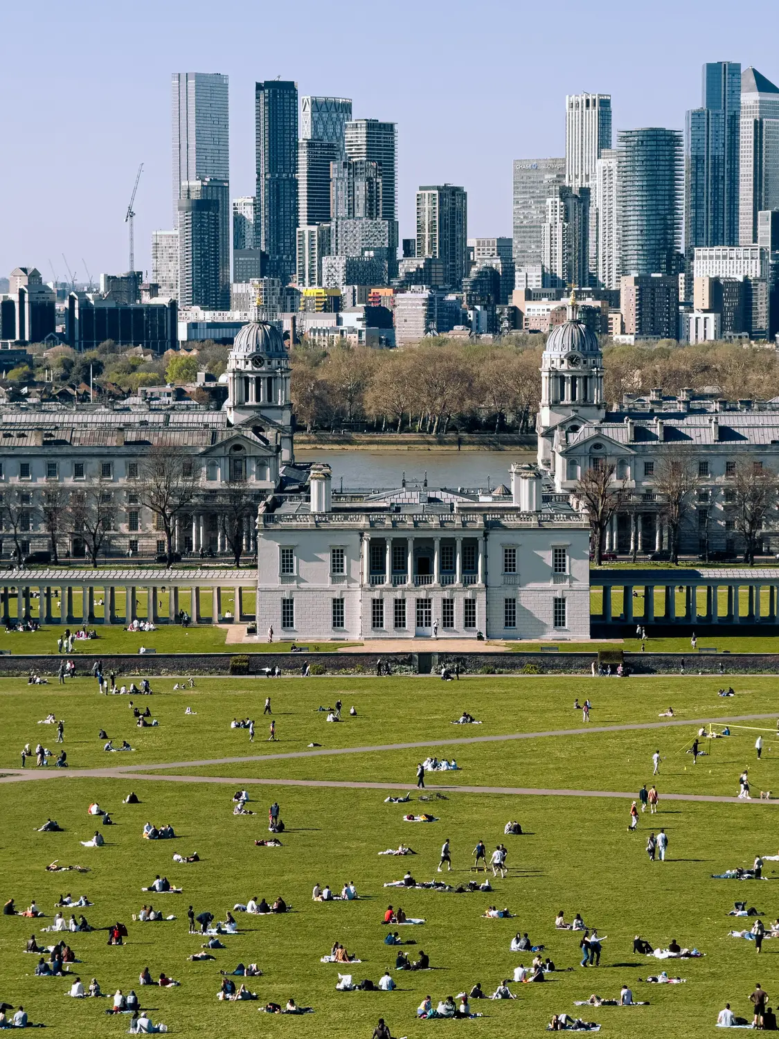 People relaxing on the wide lawns of Greenwich Park, with the Old Royal Naval College and the Canary Wharf skyline beyond.