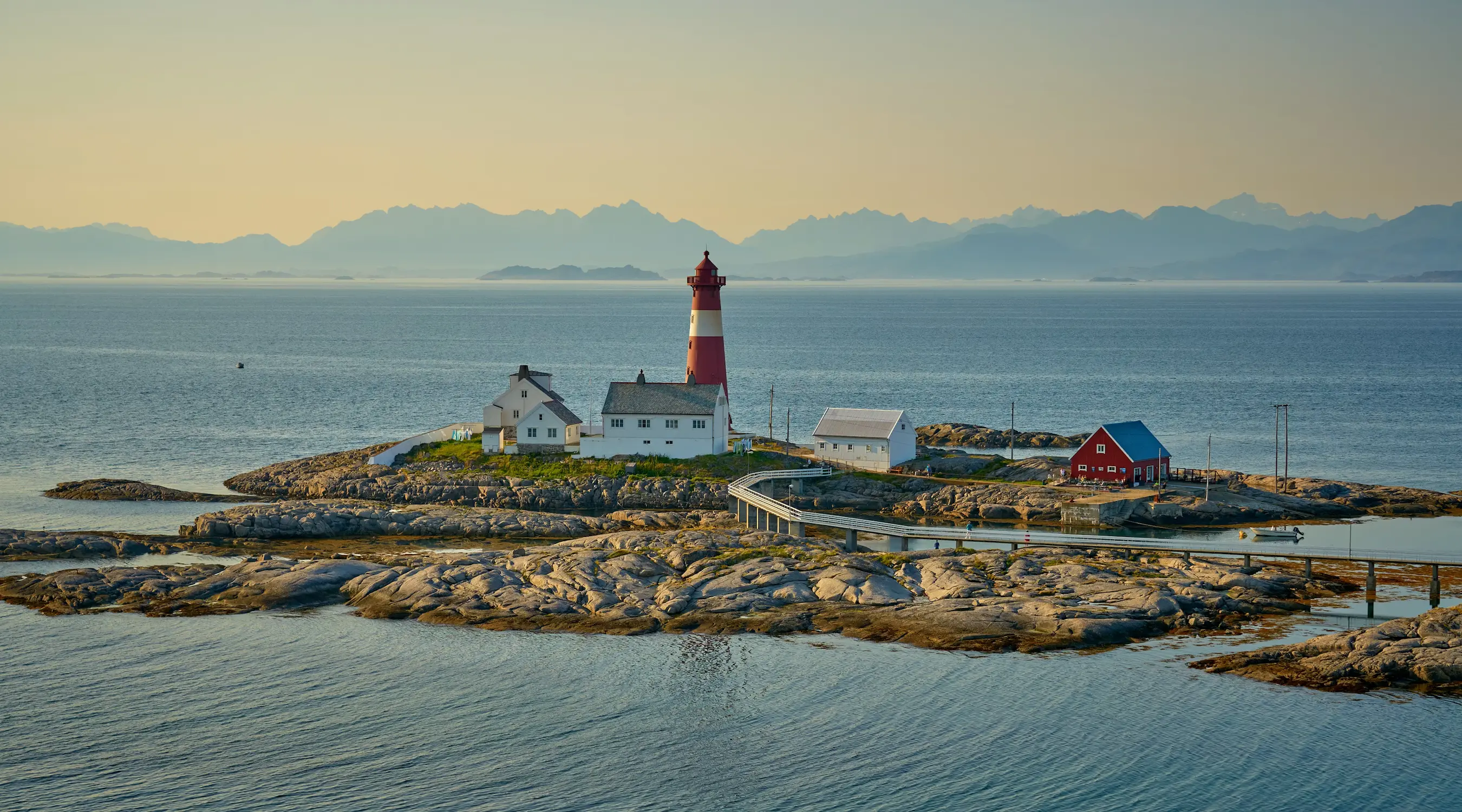 A red and white lighthouse standing on a small rocky island with a few outbuildings, distant mountains lining the hazy horizon.