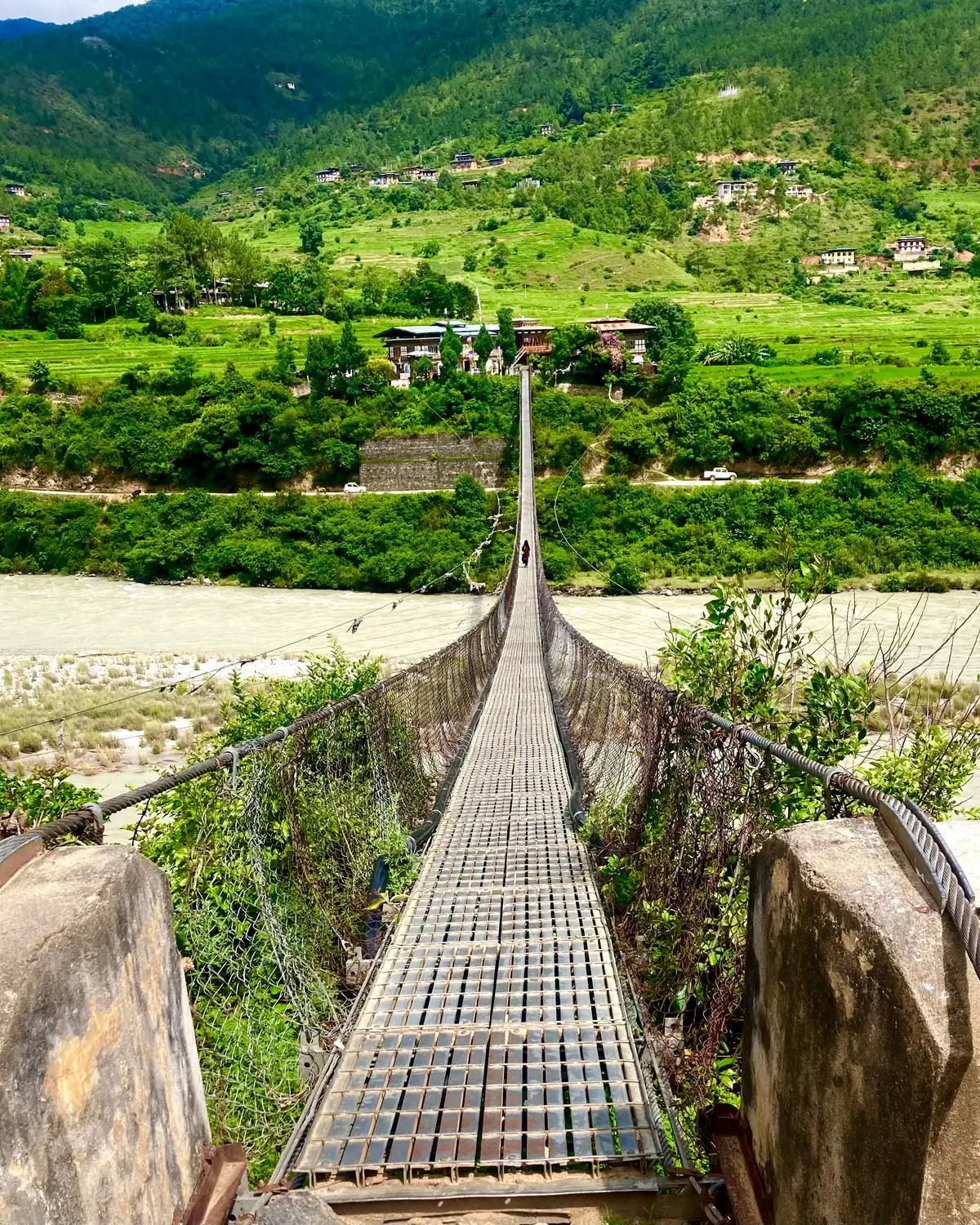 Traditional iron suspension footbridge stretching across a river valley toward lush green hills and villages in Bhutan.