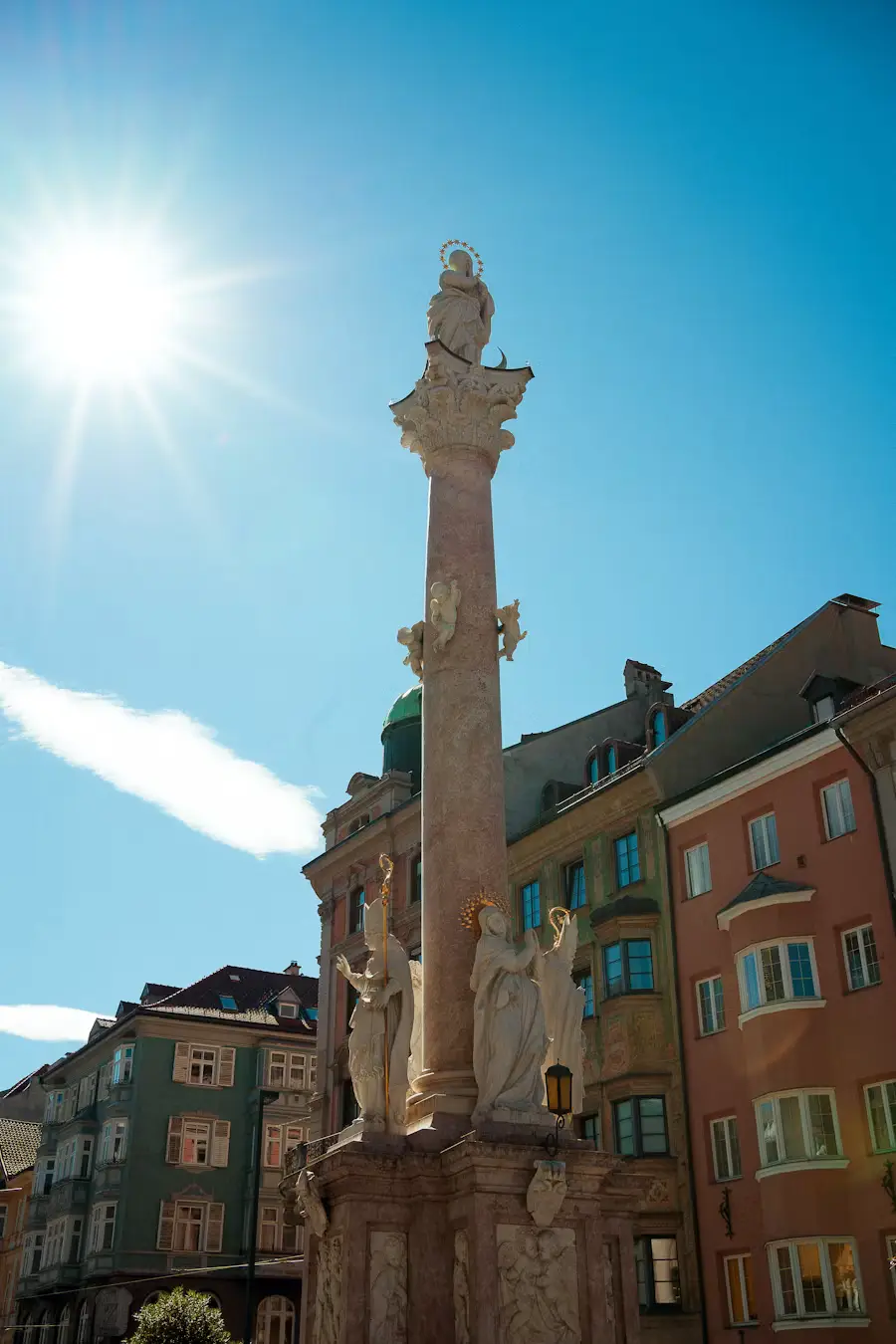 An ornate stone column monument in a plaza, backlit by bright sunlight, surrounded by traditional buildings.