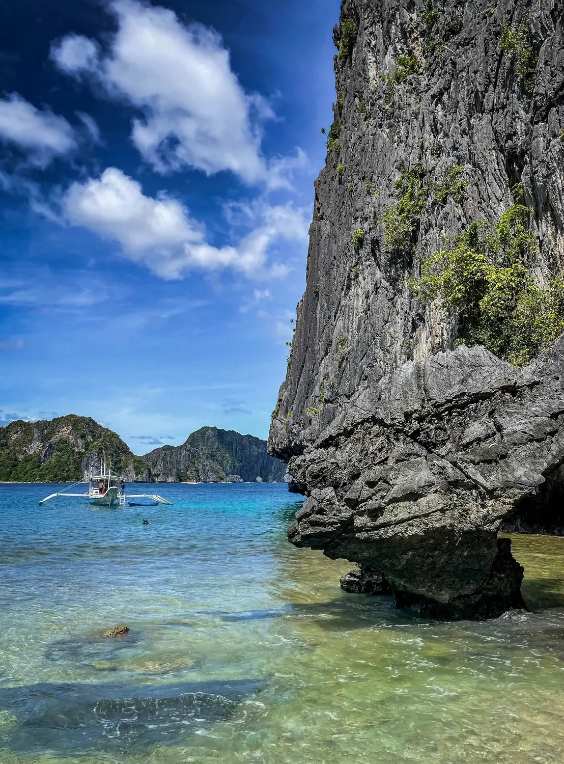 Outrigger boat on clear turquoise water beside towering limestone cliffs in El Nido, Palawan, Philippines.