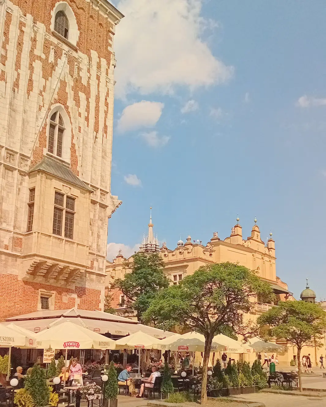 Historic European city square with a tall church tower, colorful buildings, outdoor cafés, and people.