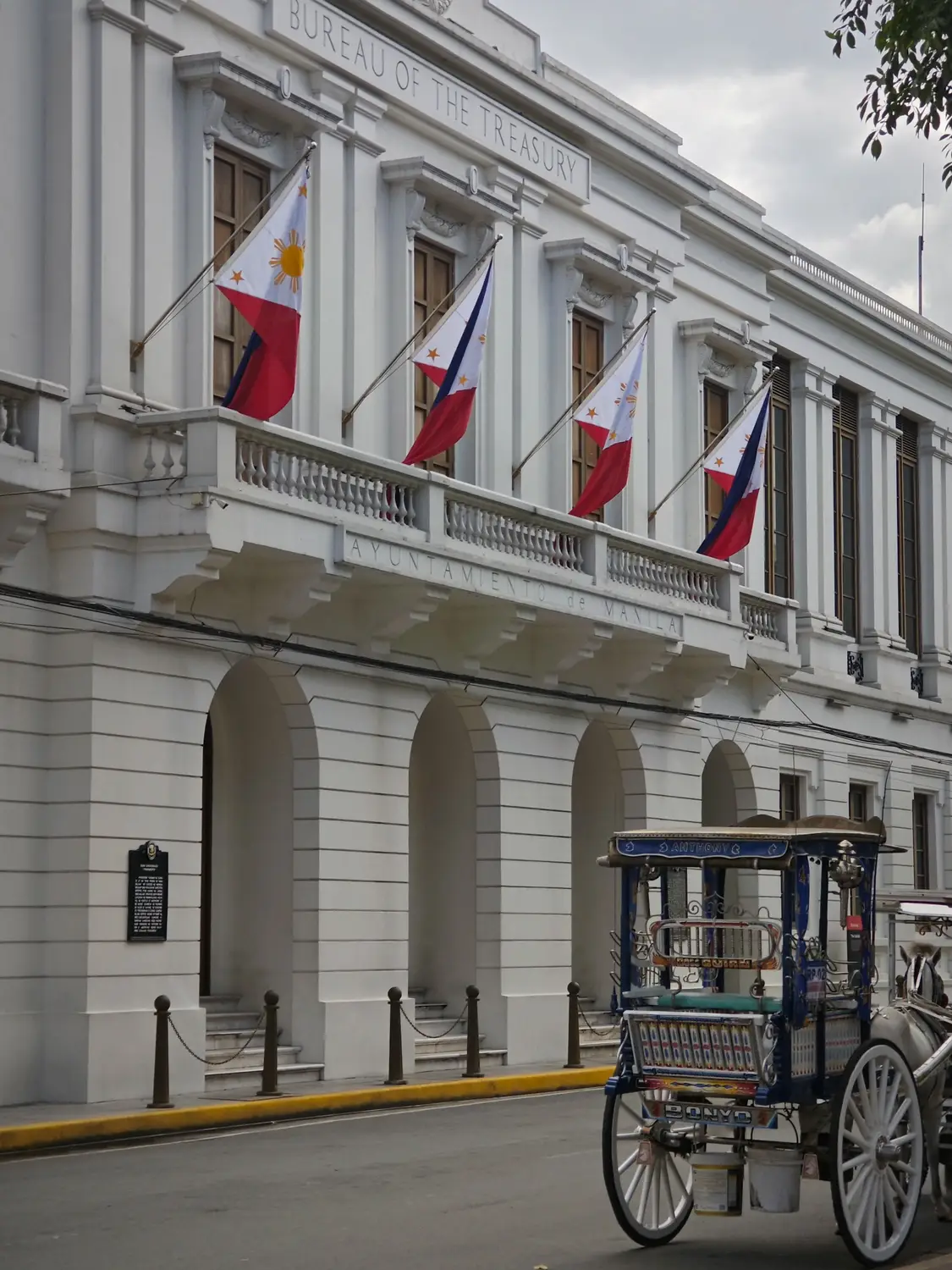 Historic white colonial Bureau of the Treasury building flying Philippine flags with a horse-drawn carriage in Intramuros, Manila, Philippines.