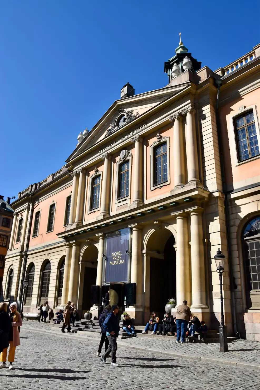 Neoclassical building facade with columned entrance, with people walking on the cobblestone square in front.