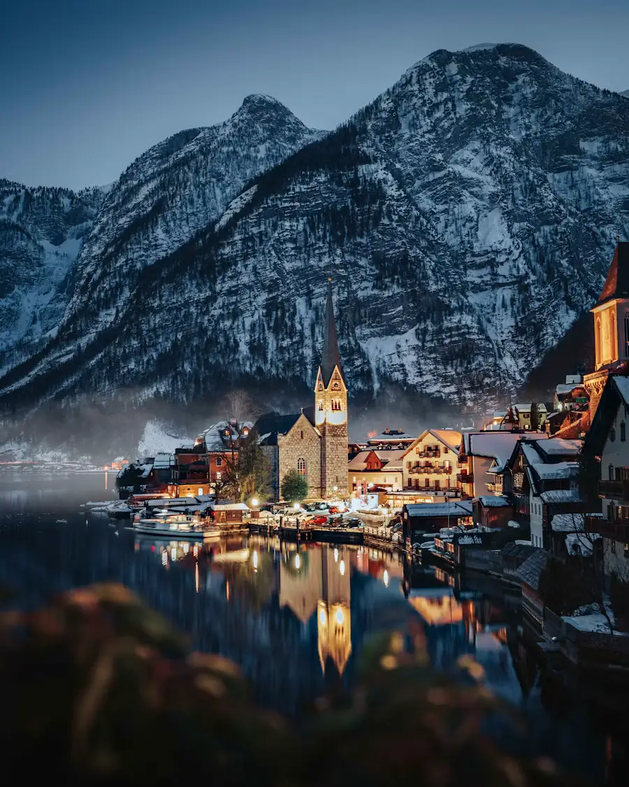 A lakeside village illuminated at dusk, with warm lights reflecting off the water beneath towering snow-covered mountains.