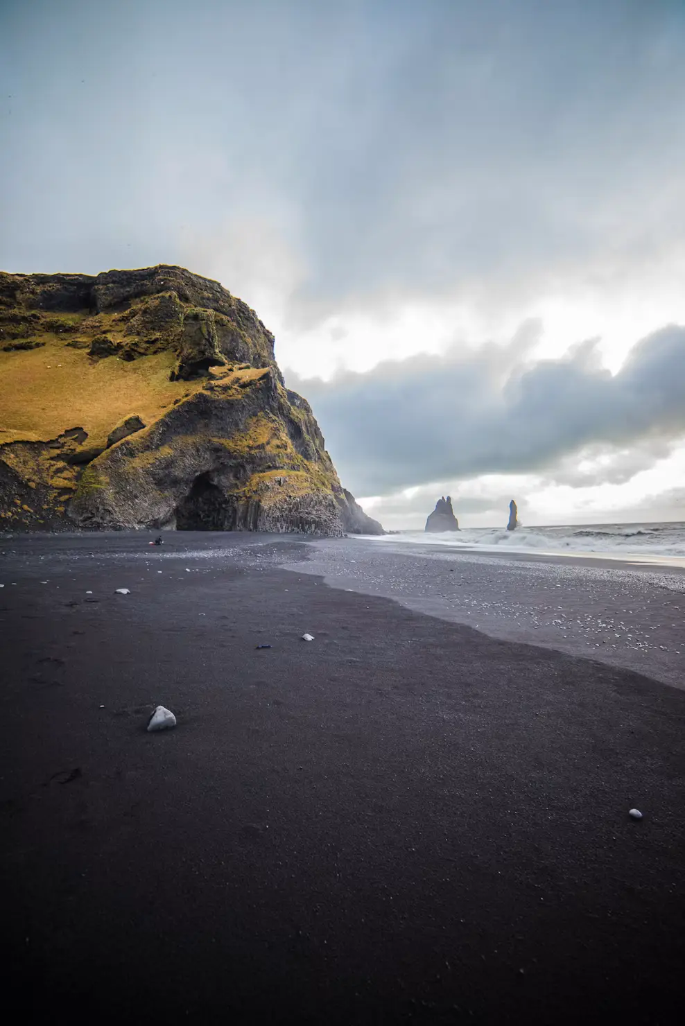 Black sand beach beneath a moss-covered cliff and cave entrance, under a hazy sky, Iceland.