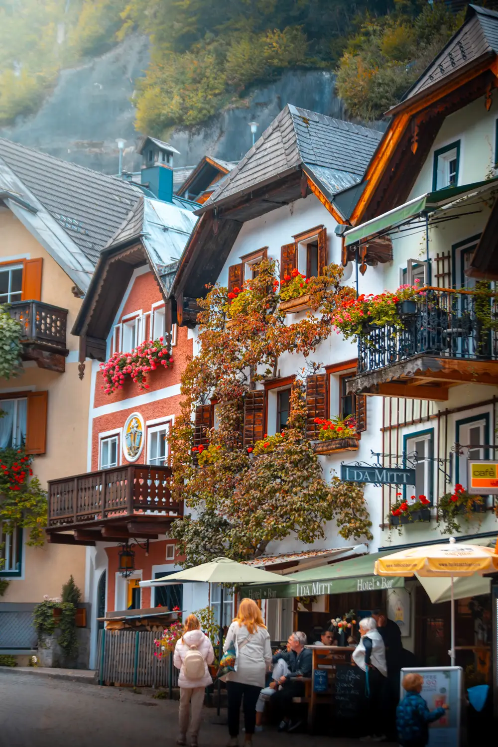 A colorful row of traditional buildings with flower-covered balconies lining a lively pedestrian street with an outdoor café.