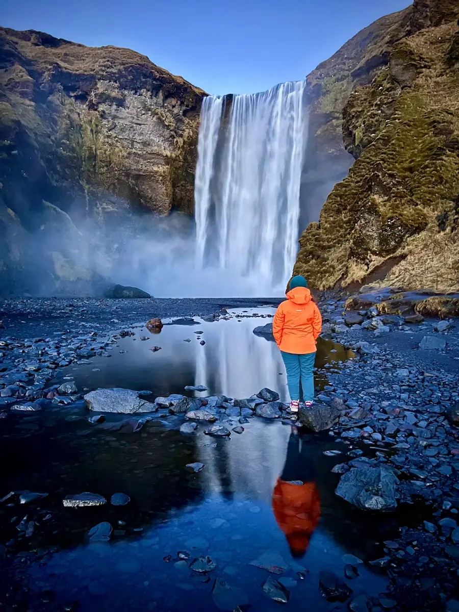 Waterfall cascading over a cliff onto a rocky riverbed, with a person in an orange jacket standing among the rocks, Iceland.