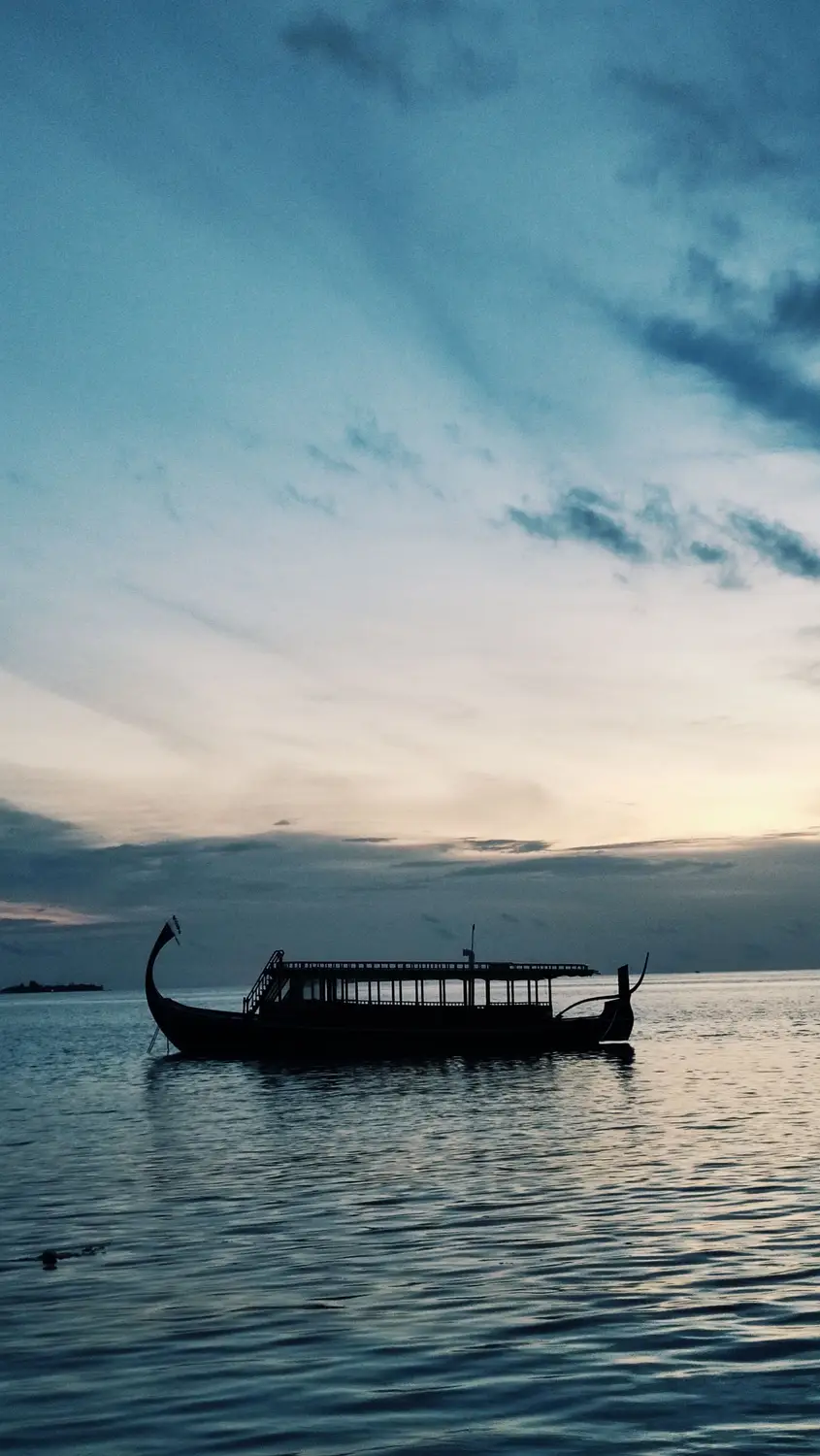 Silhouette of traditional Maldivian dhoni boat on calm ocean water at dusk with dramatic blue sunset sky