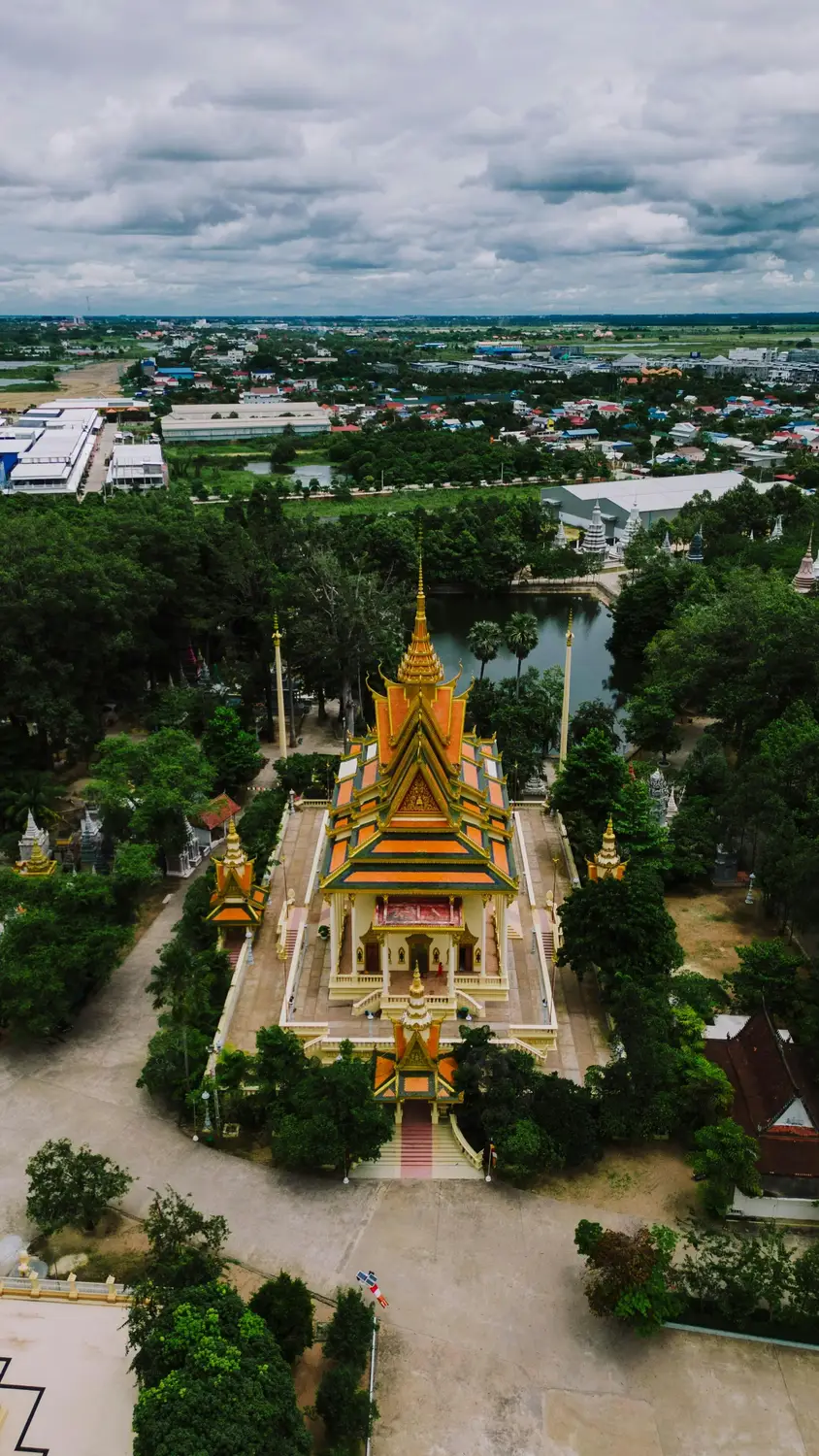 Aerial drone view of ornate golden Cambodian Buddhist temple with tiered orange roof surrounded by green trees and pond, Cambodia