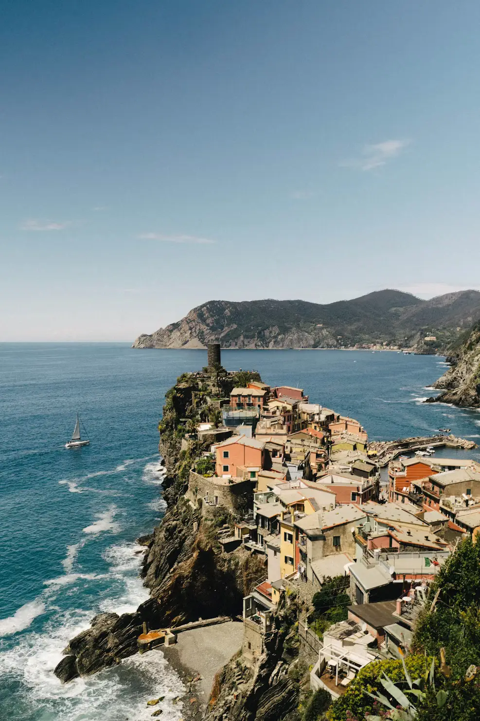 Colorful cliffside village overlooking the Mediterranean Sea with a historic stone tower, sailboat, and rugged coastline in Cinque Terre, Italy.