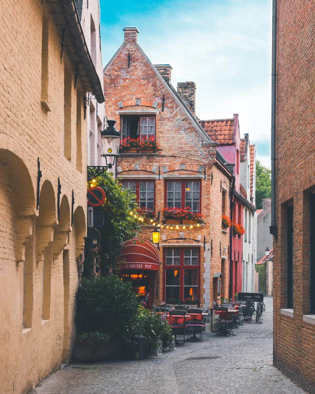 A cozy brick building with red shutters and flower boxes housing the Vlaamsche Pot restaurant on a quiet Bruges cobblestone alley.