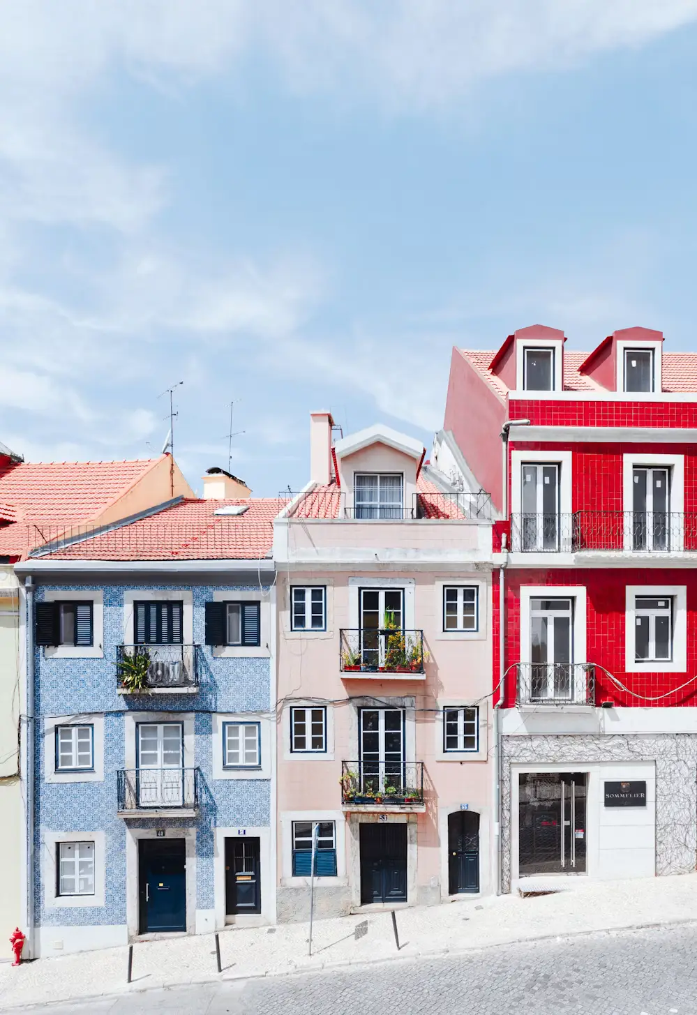 Colorful row of pastel blue, pink, and bright red houses with tiled roofs and balconies along a quiet cobblestone street.