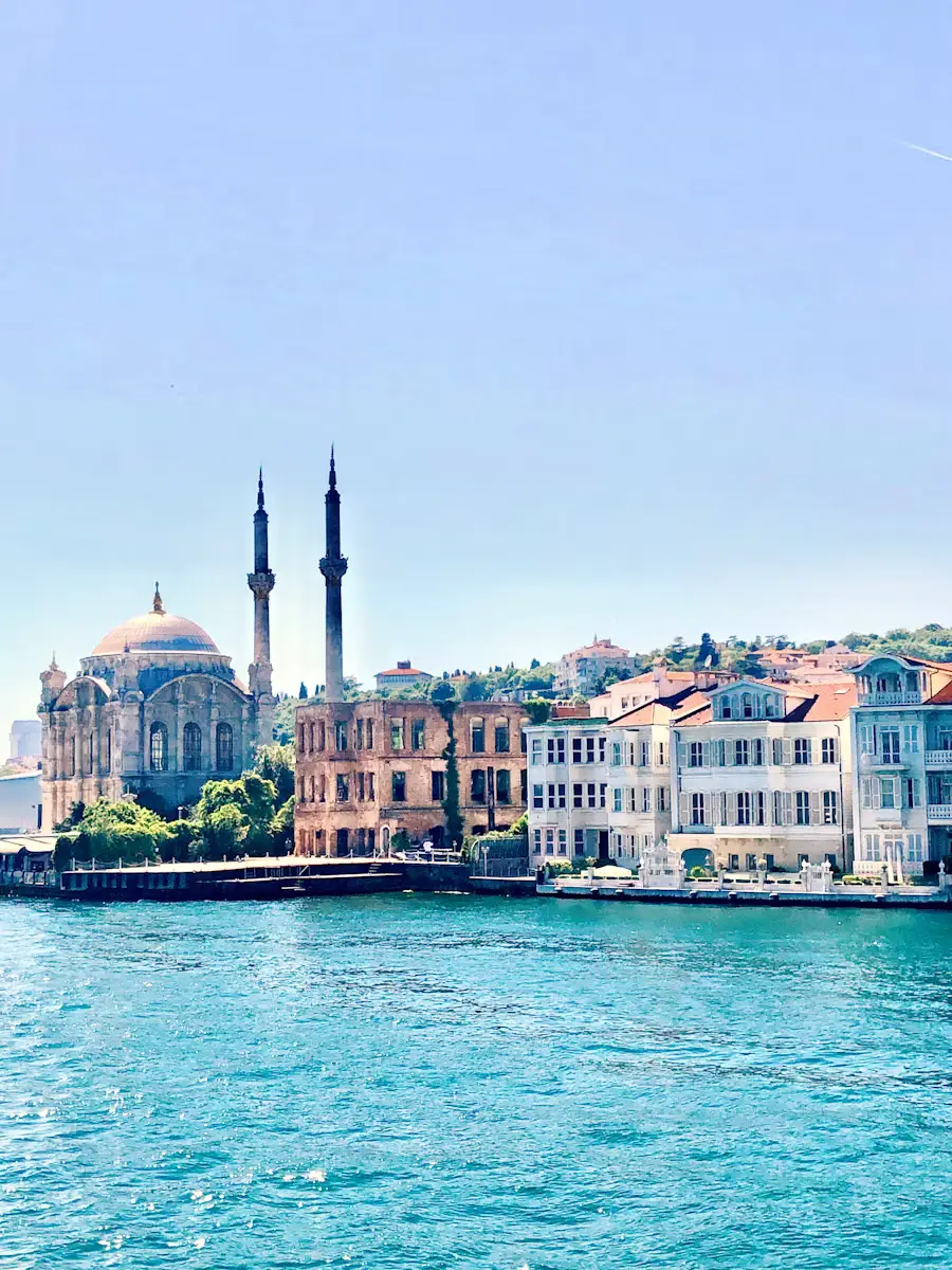 A domed mosque with twin minarets standing along the Bosphorus waterfront, with colorful buildings lining the shore.