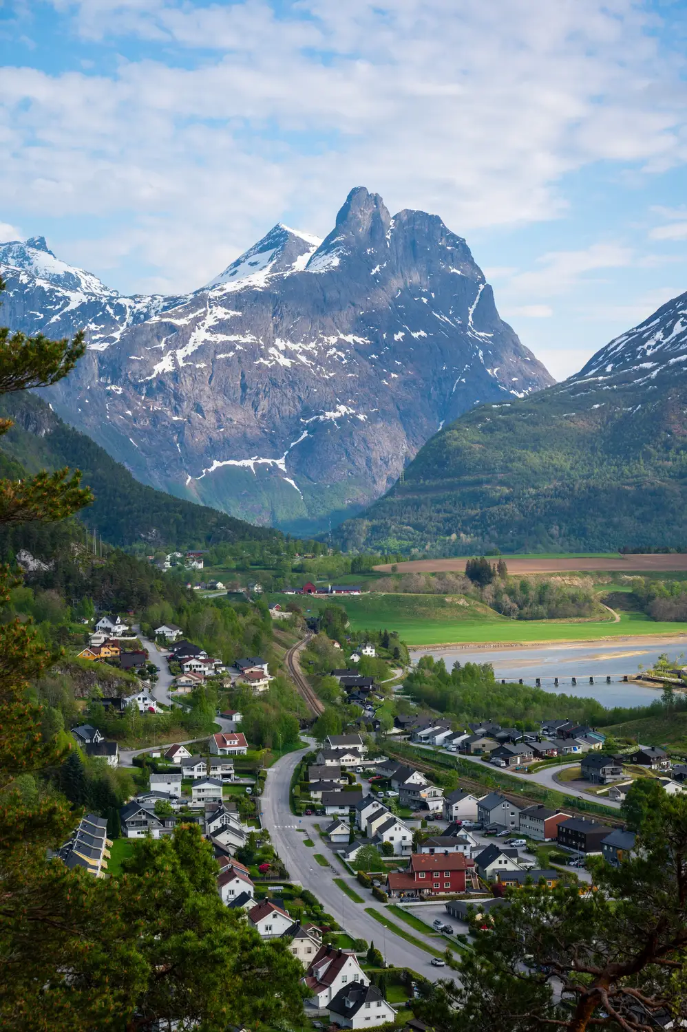 View over a valley town nestled among fields and winding roads, with jagged snow-dusted peaks towering behind.