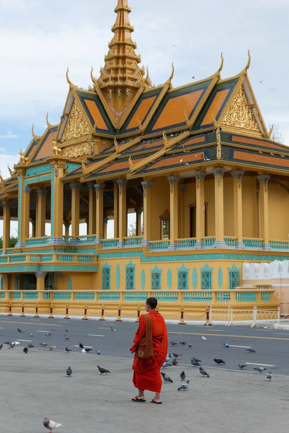 Buddhist monk in orange robe walking past pigeons in front of ornate golden Royal Palace pavilion, Phnom Penh, Cambodia