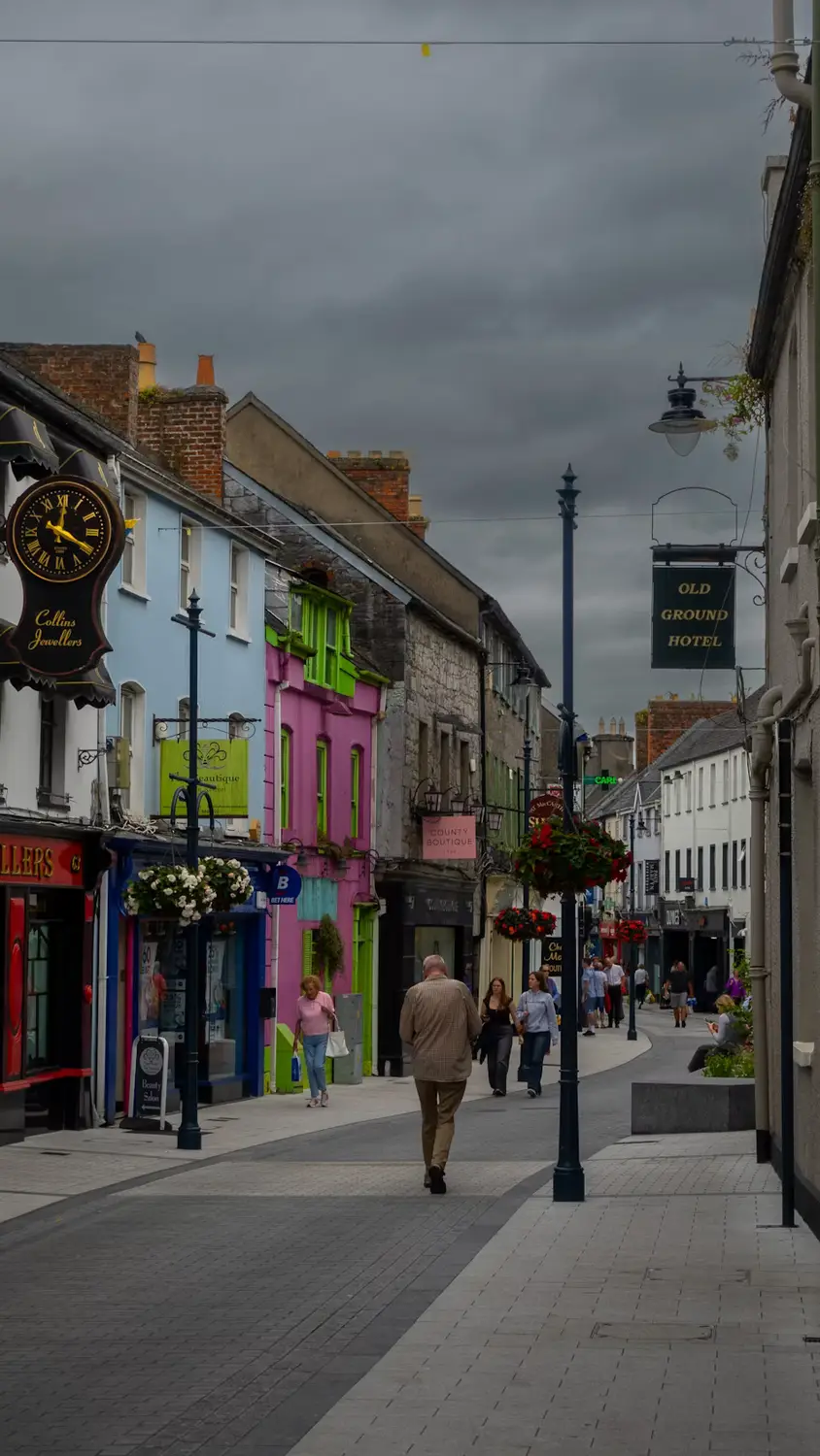 Pedestrians walking down a colorful storefront-lined street under a cloudy sky, Ireland.