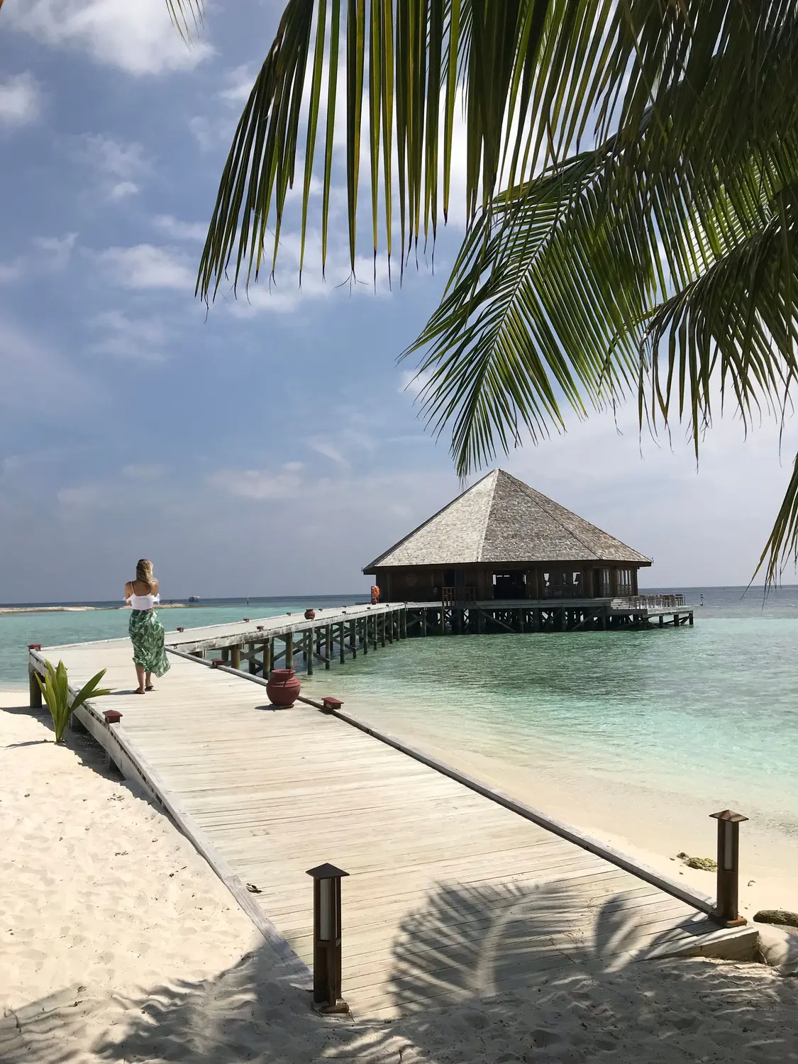 Woman walking along white sand beach with wooden jetty and thatched overwater bungalow and calm turquoise ocean in Maldives