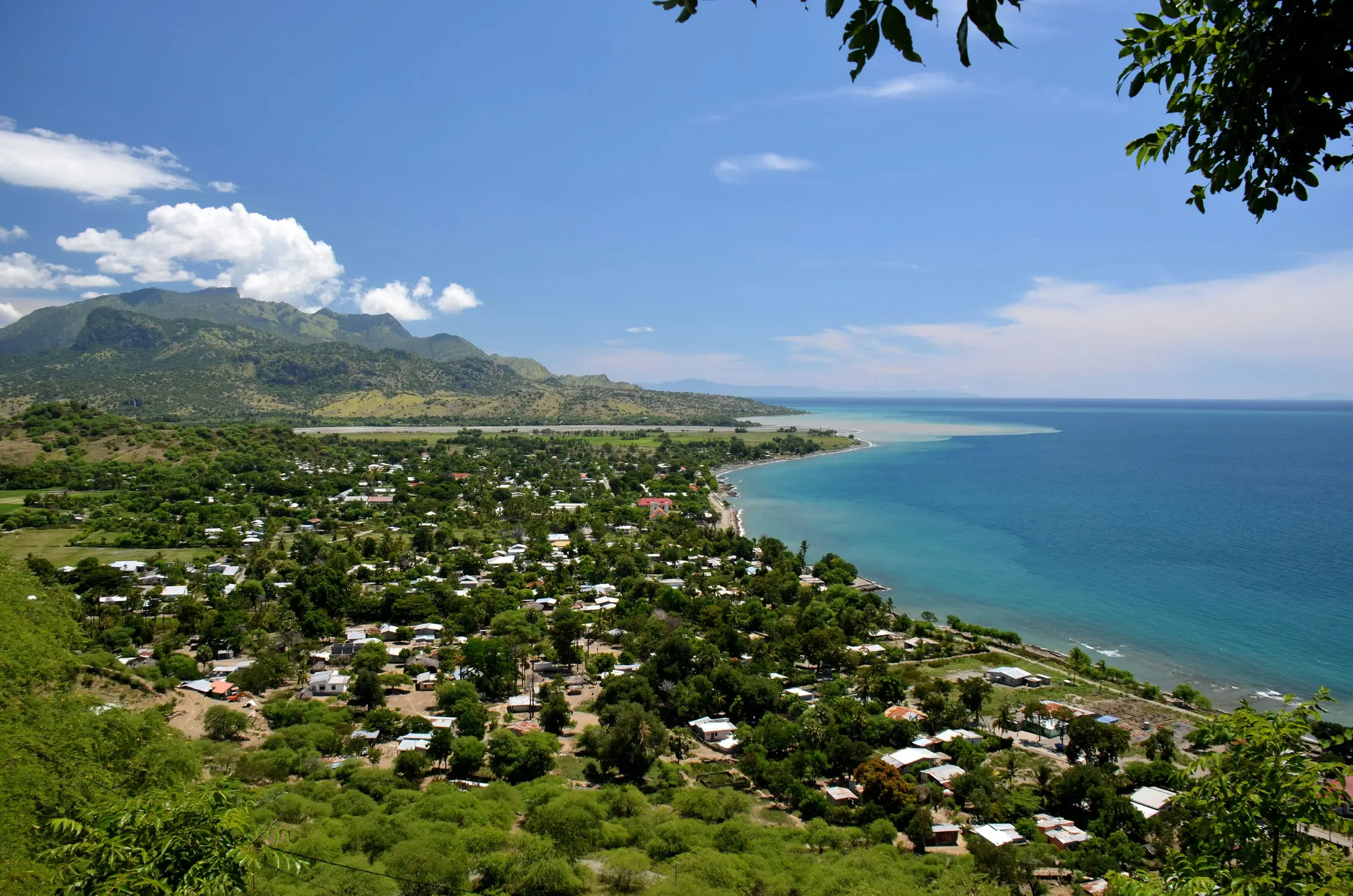 Coastal town in Timor-Leste with lush green hills, scattered houses, and a curved turquoise shoreline under a clear blue sky.