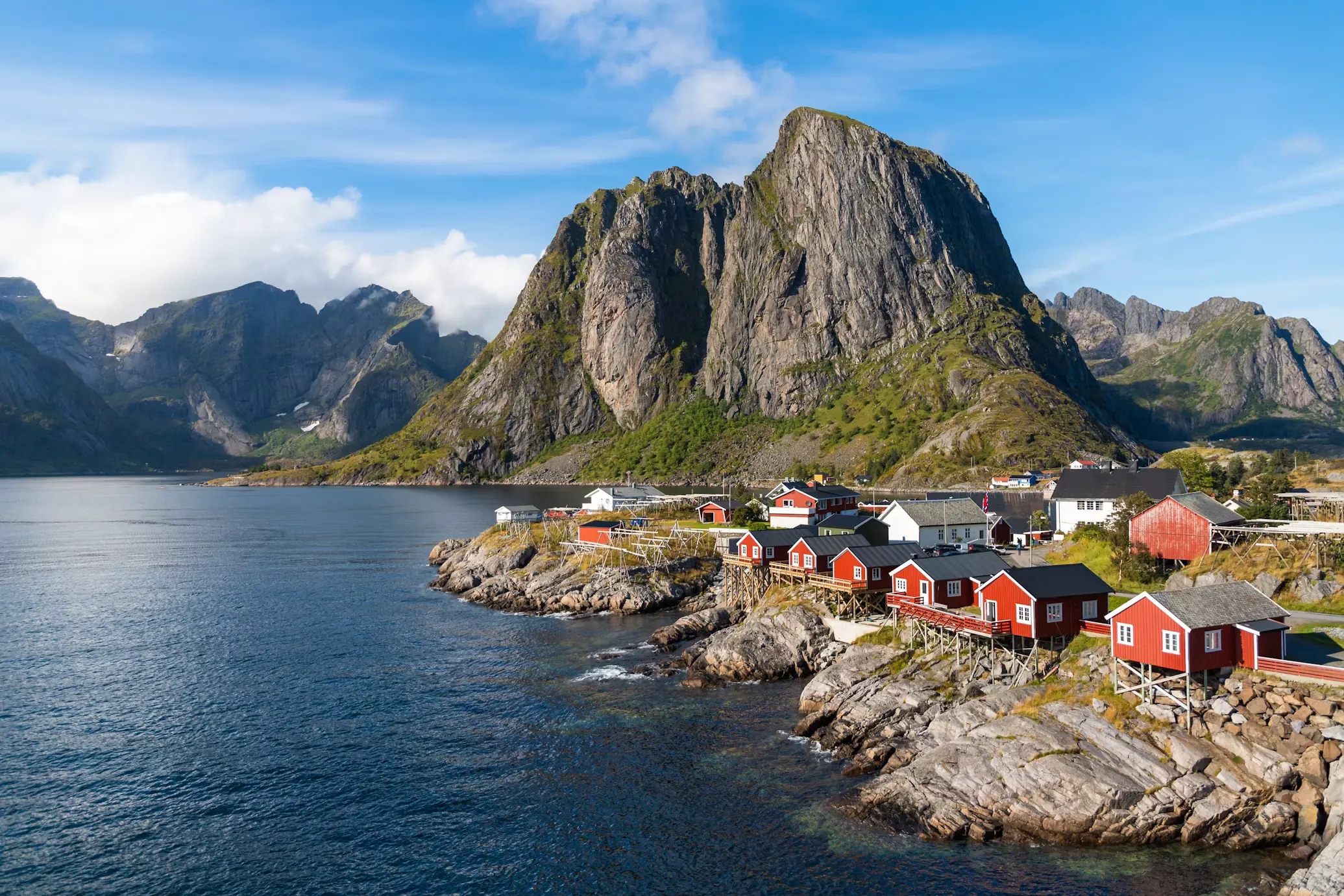 Red fishing cabins perched on the rocky shoreline of a Lofoten village, with steep mountains rising across the calm water.