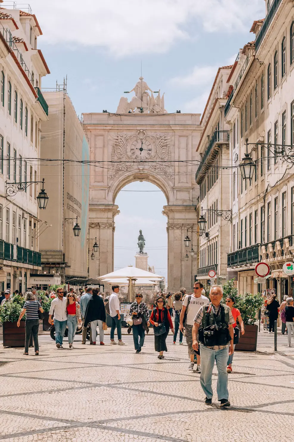 Crowded pedestrian street leading to a grand triumphal arch with a clock, flanked by historic buildings and ornate street lamps.