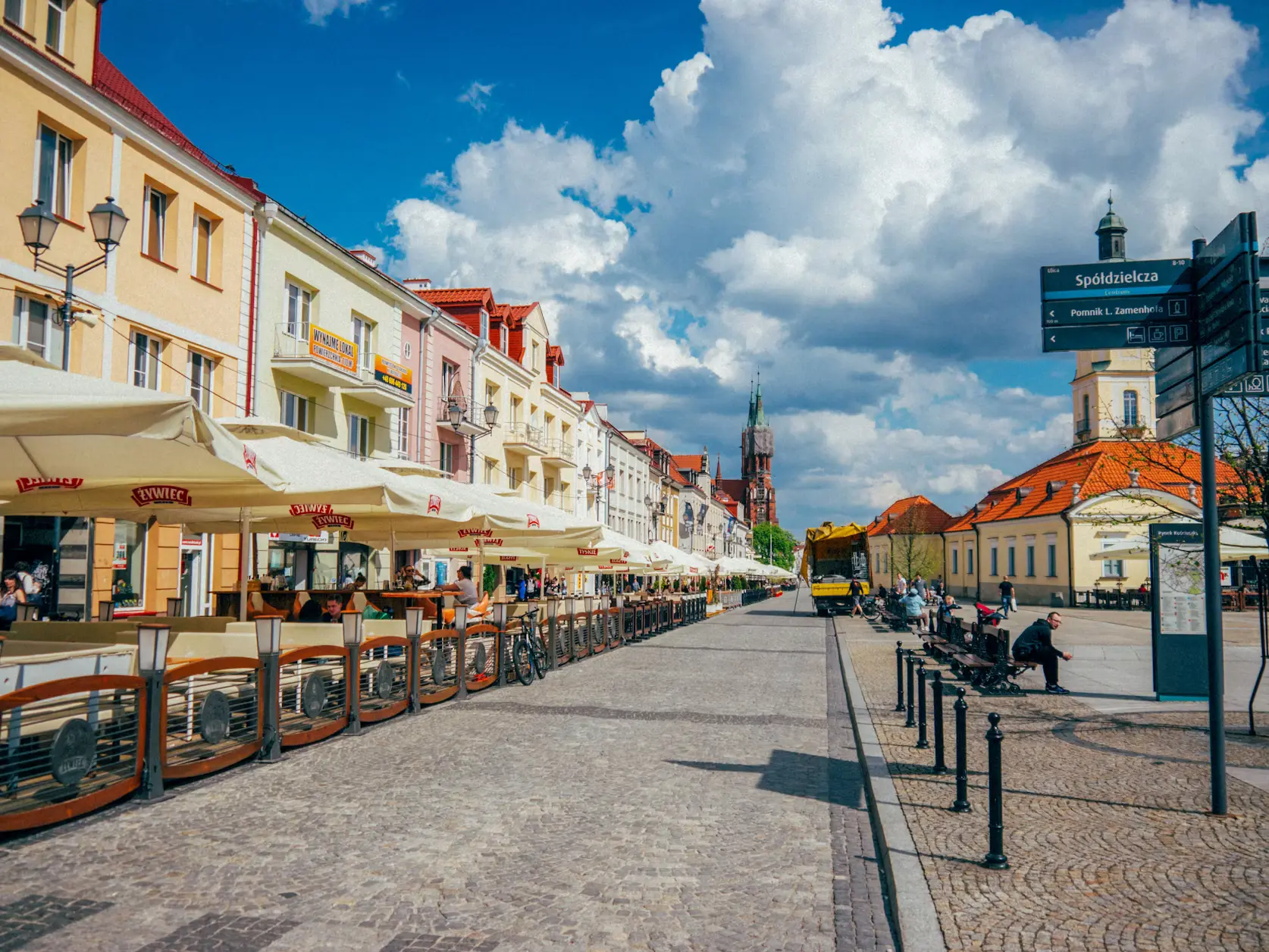 Cobblestone pedestrian street lined with pastel-colored buildings and outdoor cafés, tram tracks running down the center, Poland.