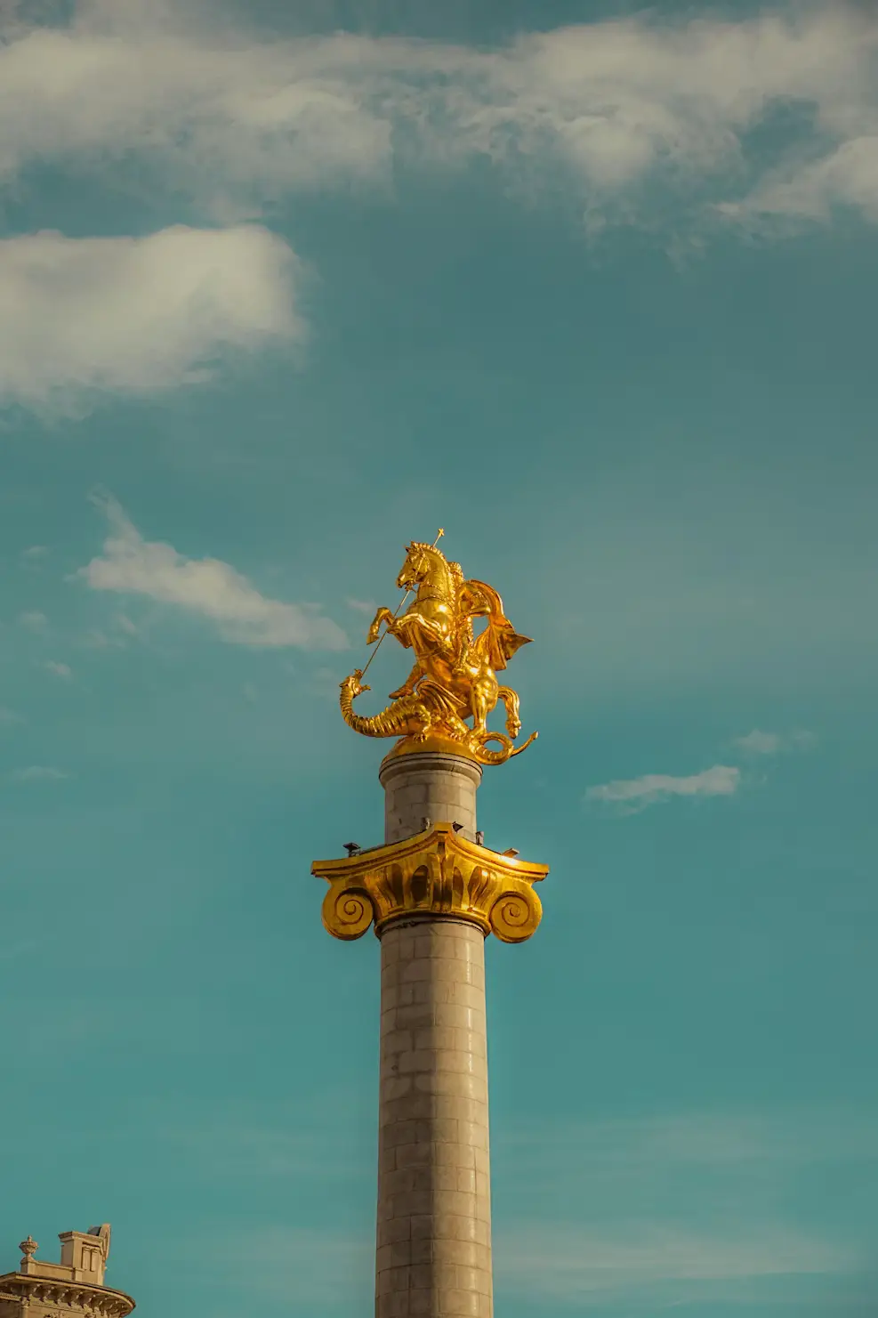 Golden equestrian statue atop a tall stone column against a blue sky with scattered clouds.