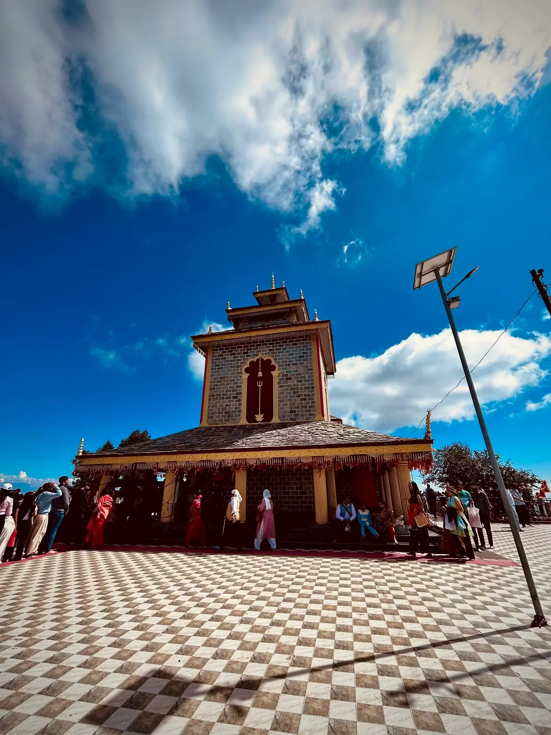 Hindu hilltop temple with trishul symbol and checkered marble courtyard with pilgrims under blue sky, Uttarakhand, India