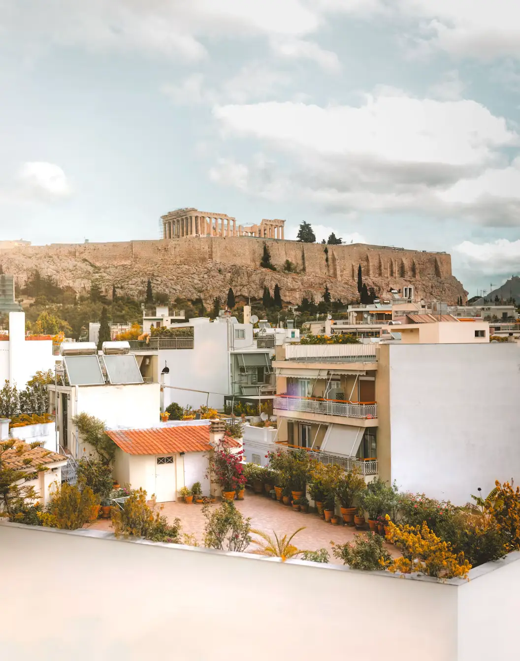Rooftop view of Athens apartment buildings with greenery, overlooking the Acropolis in the distance under a cloudy sky.