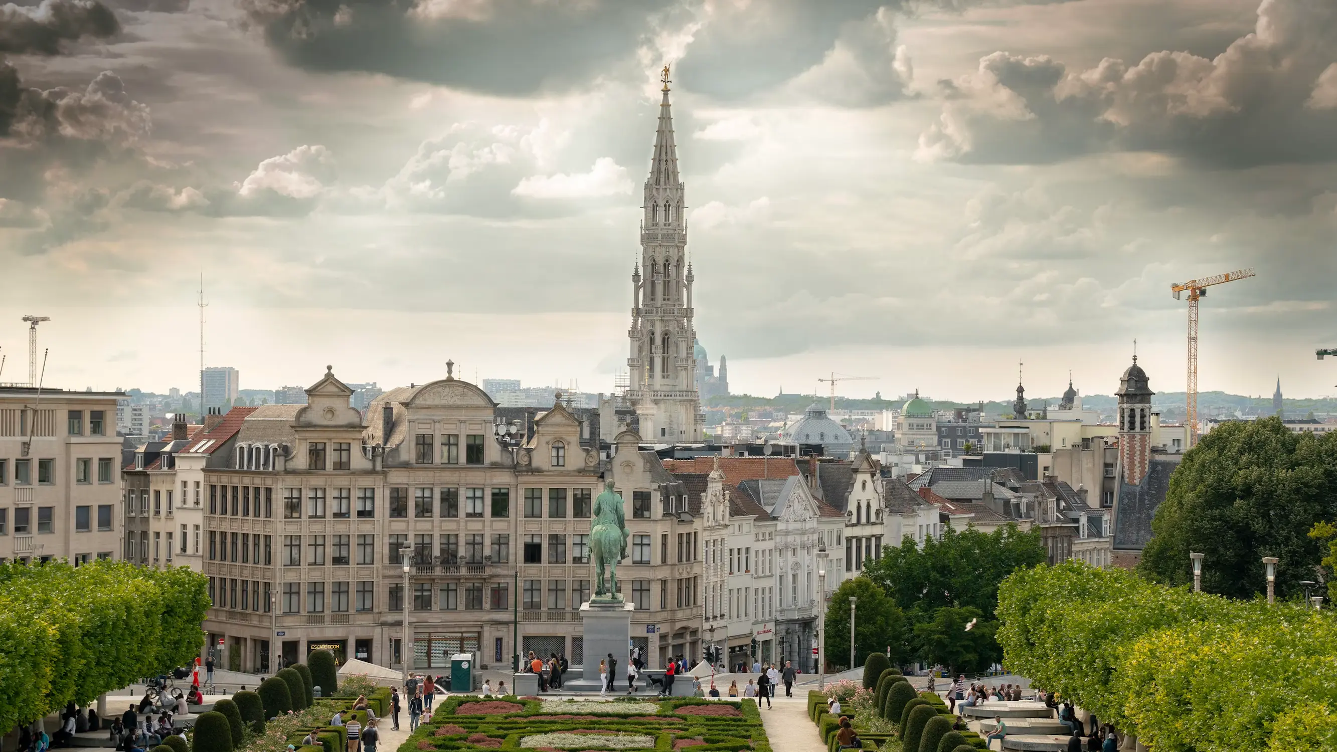 Panoramic view of Brussels' skyline from the Mont des Arts gardens, with Town Hall's spire rising above rooftops and a bronze equestrian statue in the foreground.