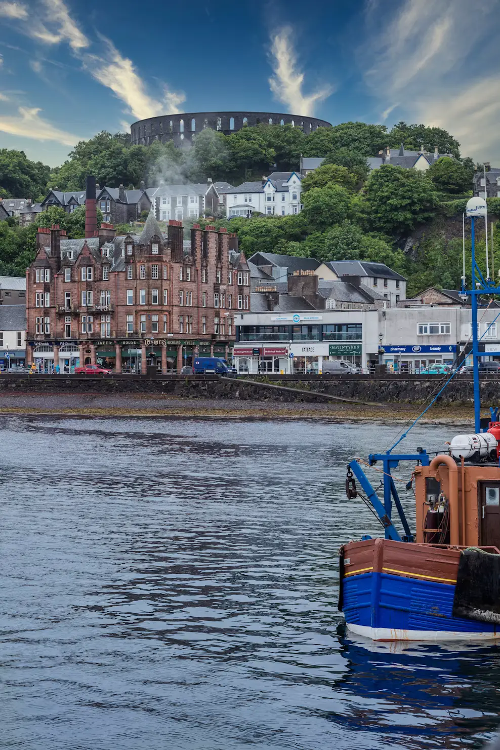 A blue fishing boat in Oban harbour, with waterfront buildings and McCaig's Tower on the hillside under a dramatic sky.