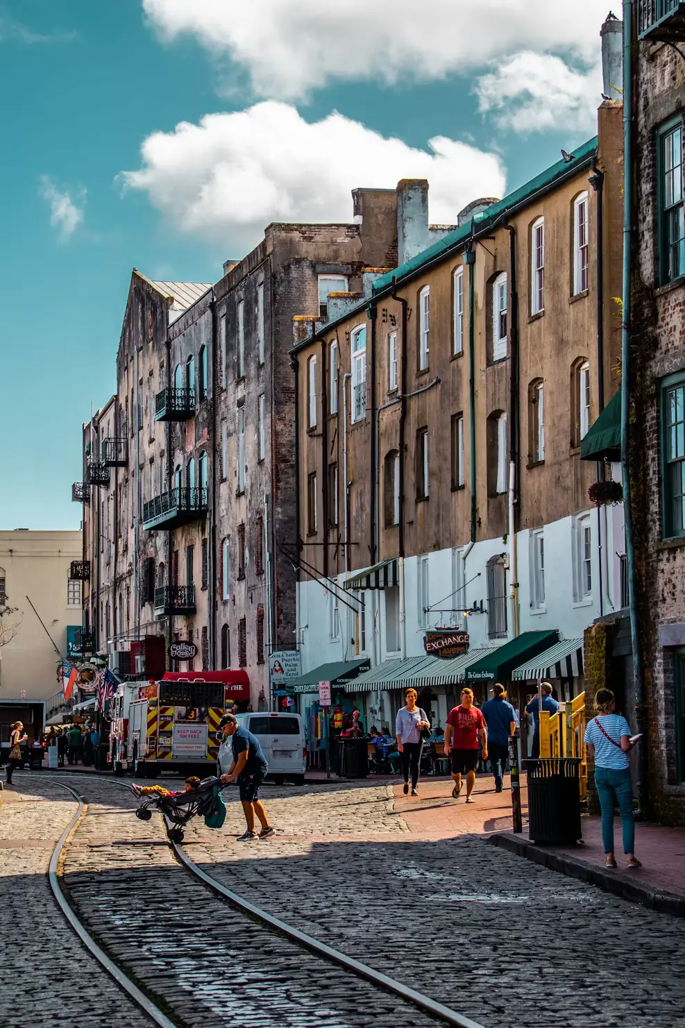 Cobblestone street lined with historic buildings, shops, and streetcar tracks under a bright blue sky.